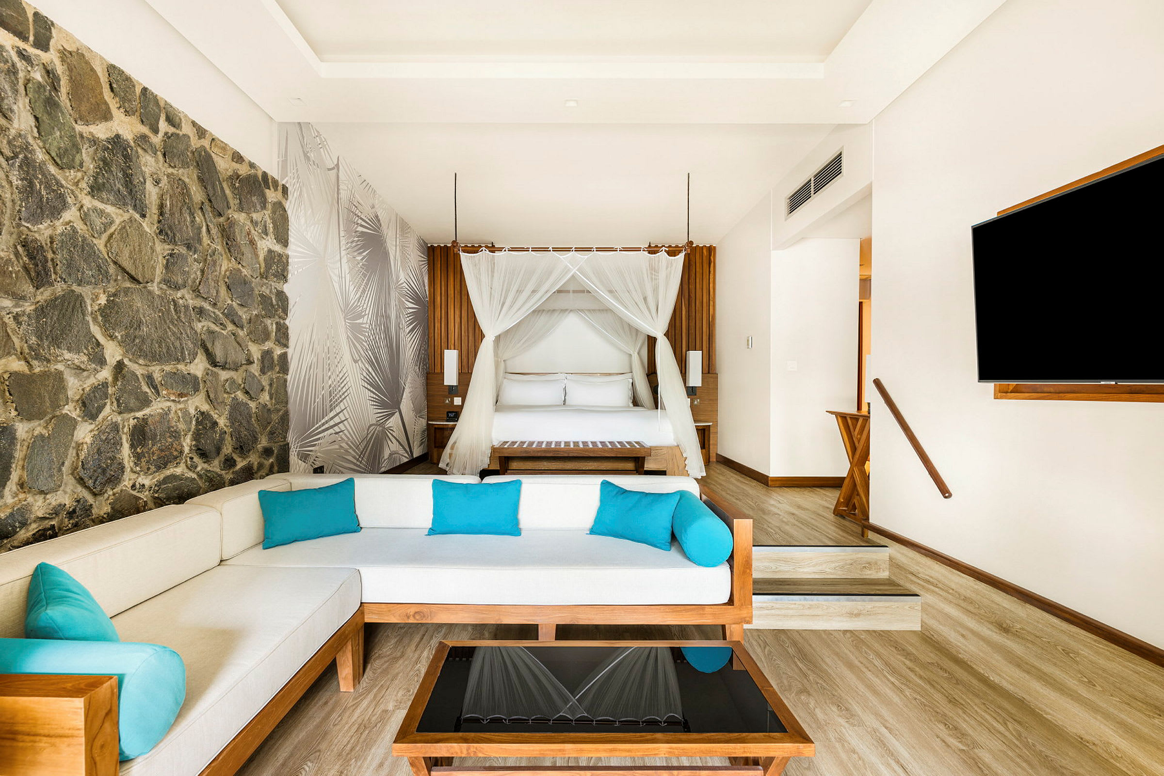 Ett modernt hotellrum med en dubbelsäng med baldakin, en vit soffa med blå kuddar och ett soffbord. En stenvägg dekorerar ena sidan av rummet. (AI-genererad text)