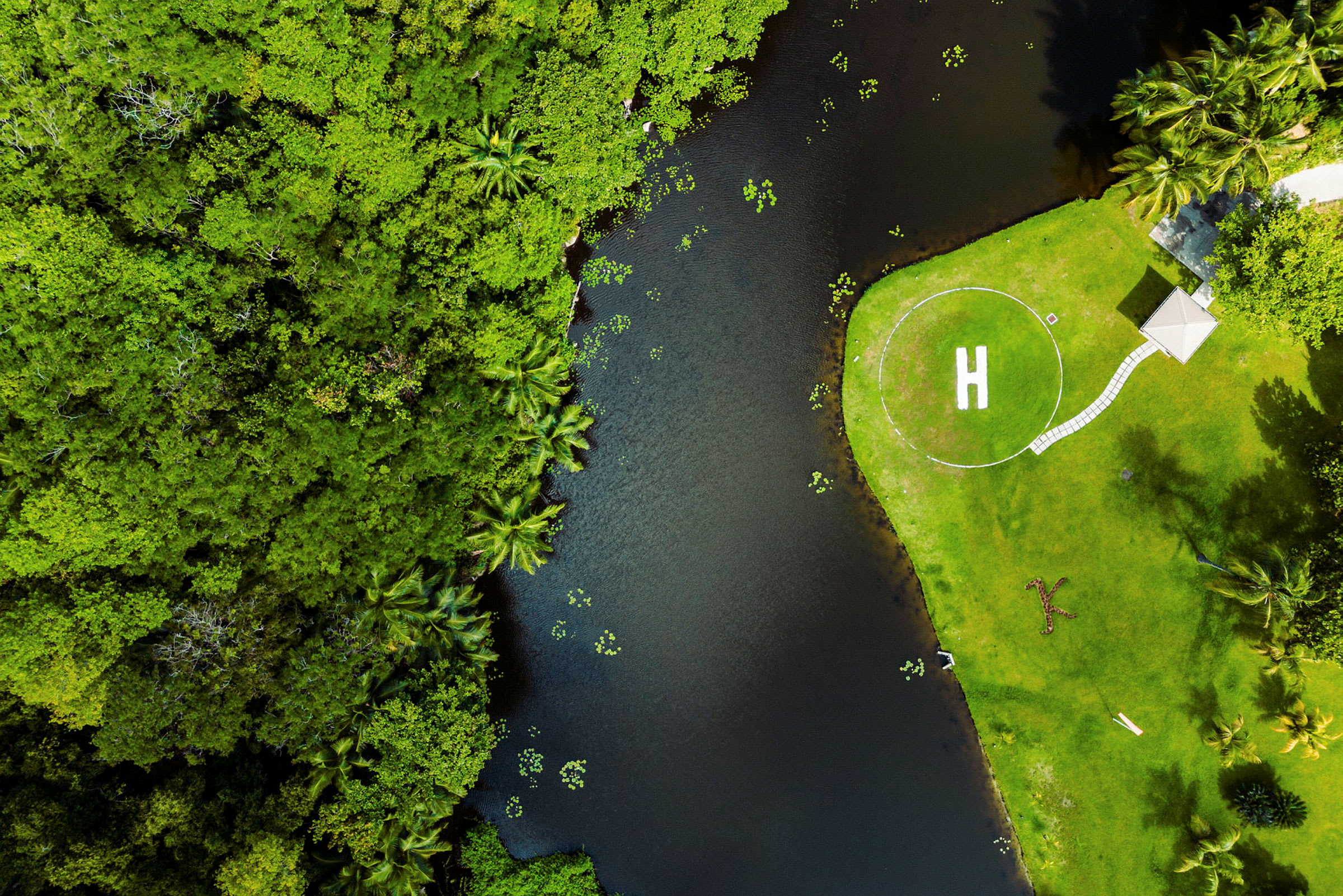 Flygfoto av ett grönskande område med en helikopterplatta markerad med ett "H" bredvid en liten vattensamling. (AI-genererad text)