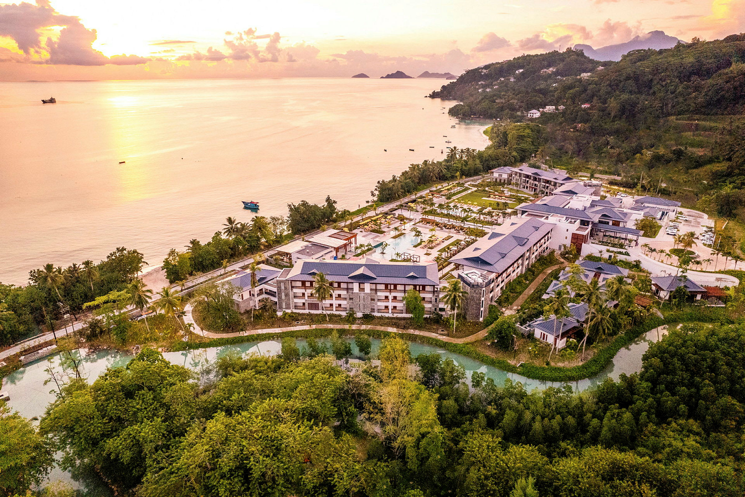 Flygbild över ett hotellkomplex omgivet av natur och beläget nära en kust med strand och hav vid solnedgång. (AI-genererad text)