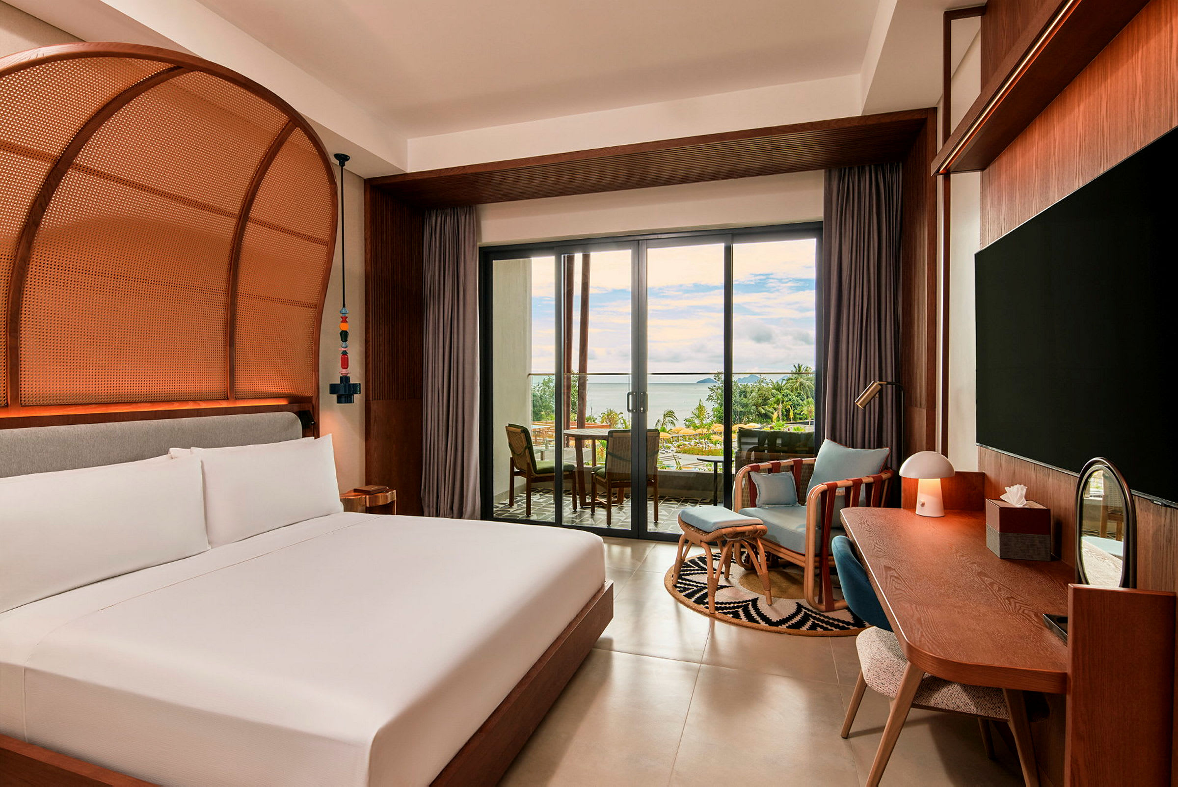 Ett hotellrum med en stor säng, skrivbord och sittgrupp bredvid fönsterdörrar som vetter mot en terrass med utsikt över havet. (AI-genererad text)