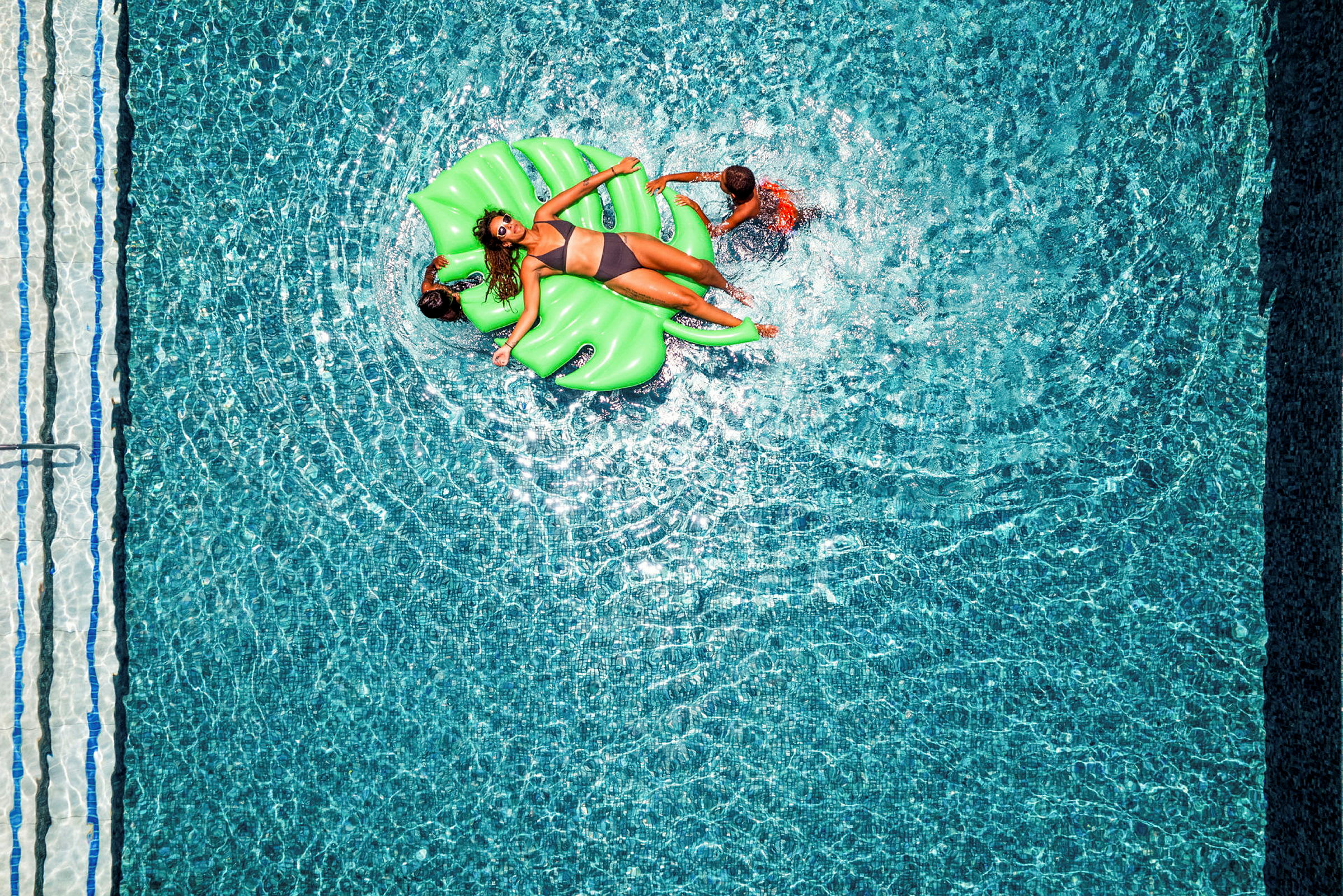 En person ligger på en grön flytmadrass i en pool medan två andra personer leker i vattnet. (AI-genererad text)