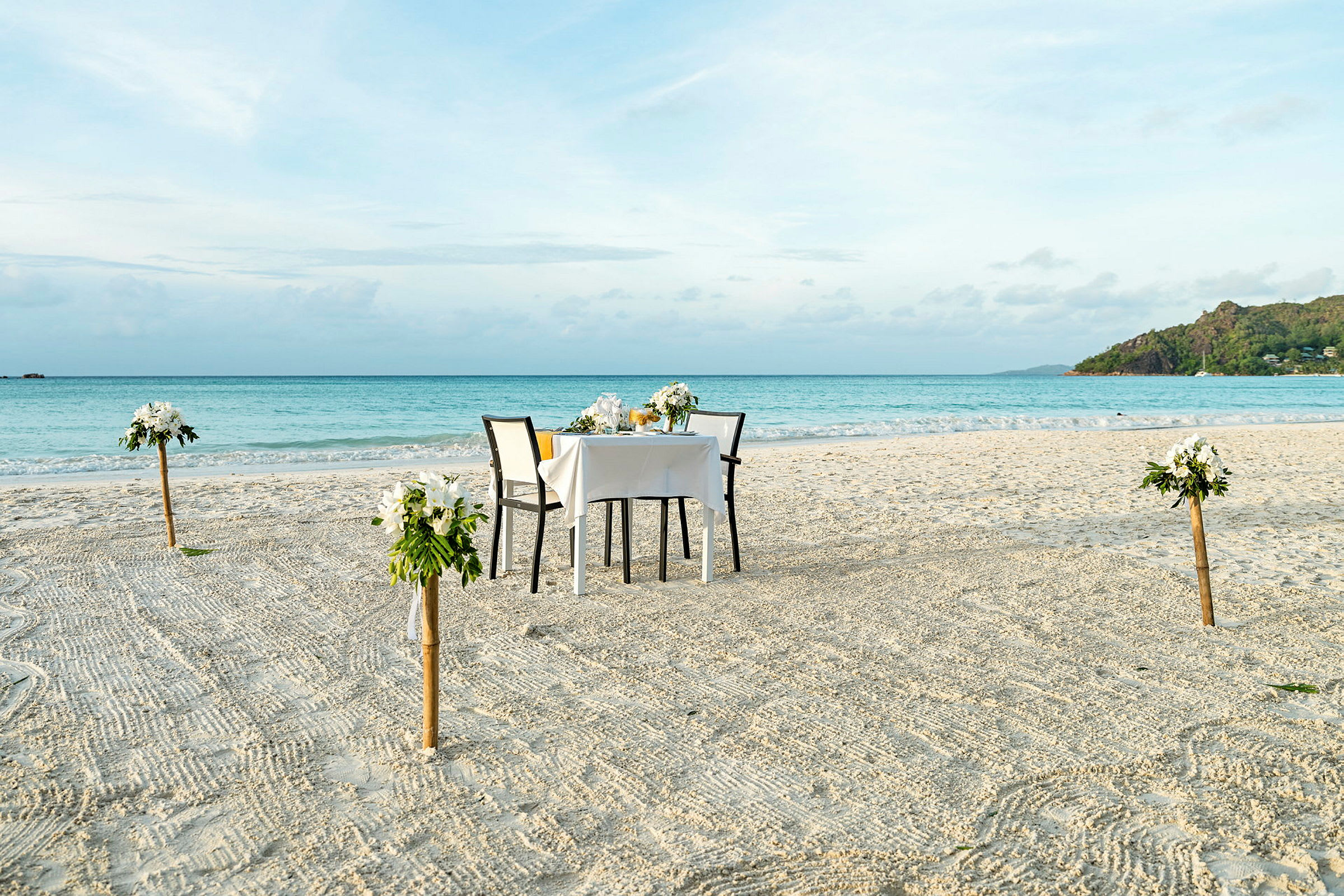 Ett dukat bord med två stolar står på en sandstrand. Bordet är dekorerat med blomsterarrangemang, och fyra blomsterdekorationer finns på stolpar kring bordet. I bakgrunden syns havet och en grön klippa. (AI-genererad text)