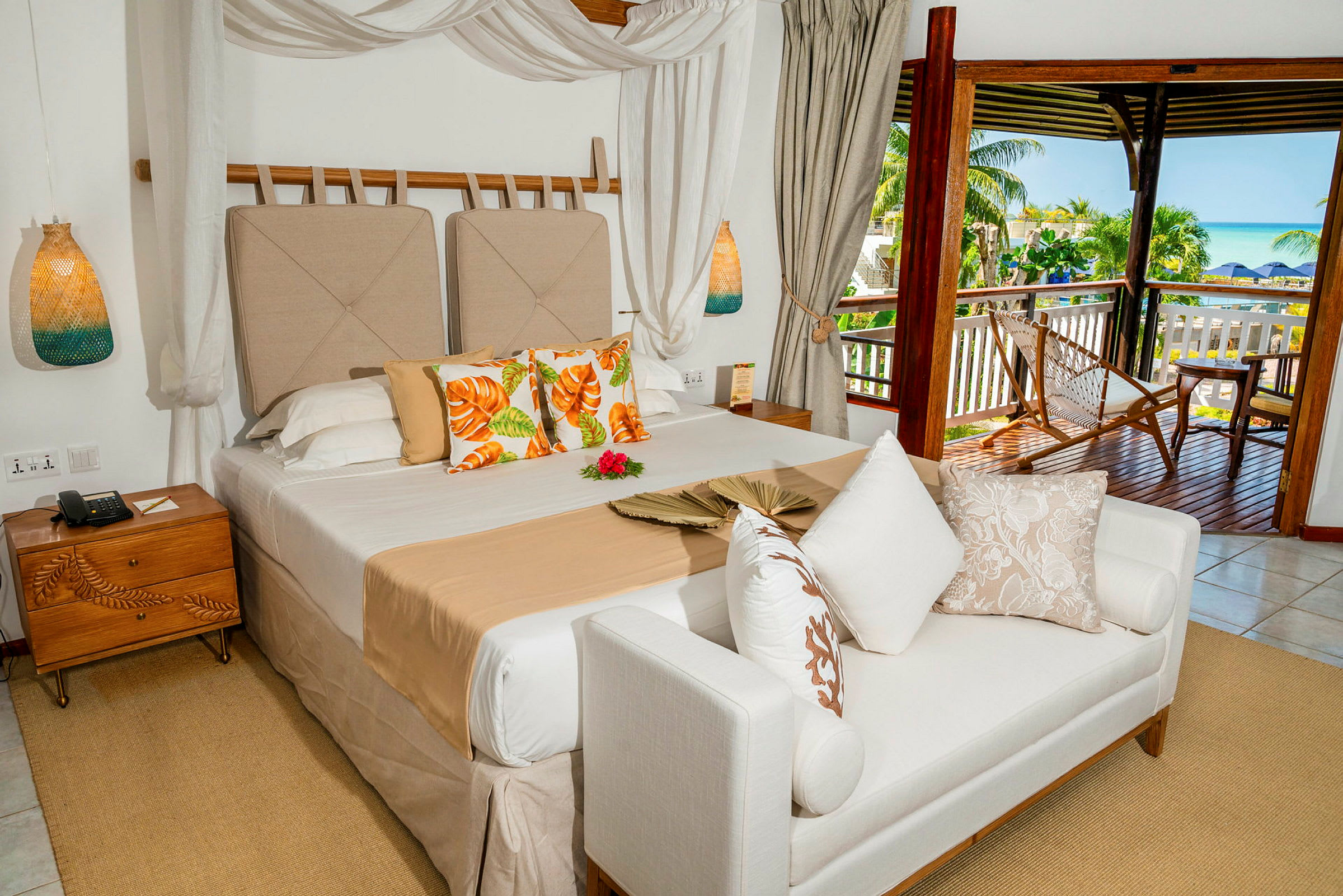 Ett hotellrum med en säng med beige sängkläder, dekorerade kuddar, och en vit soffa framför. I bakgrunden syns en balkong med utsikt över tropisk växtlighet och havet. (AI-genererad text)