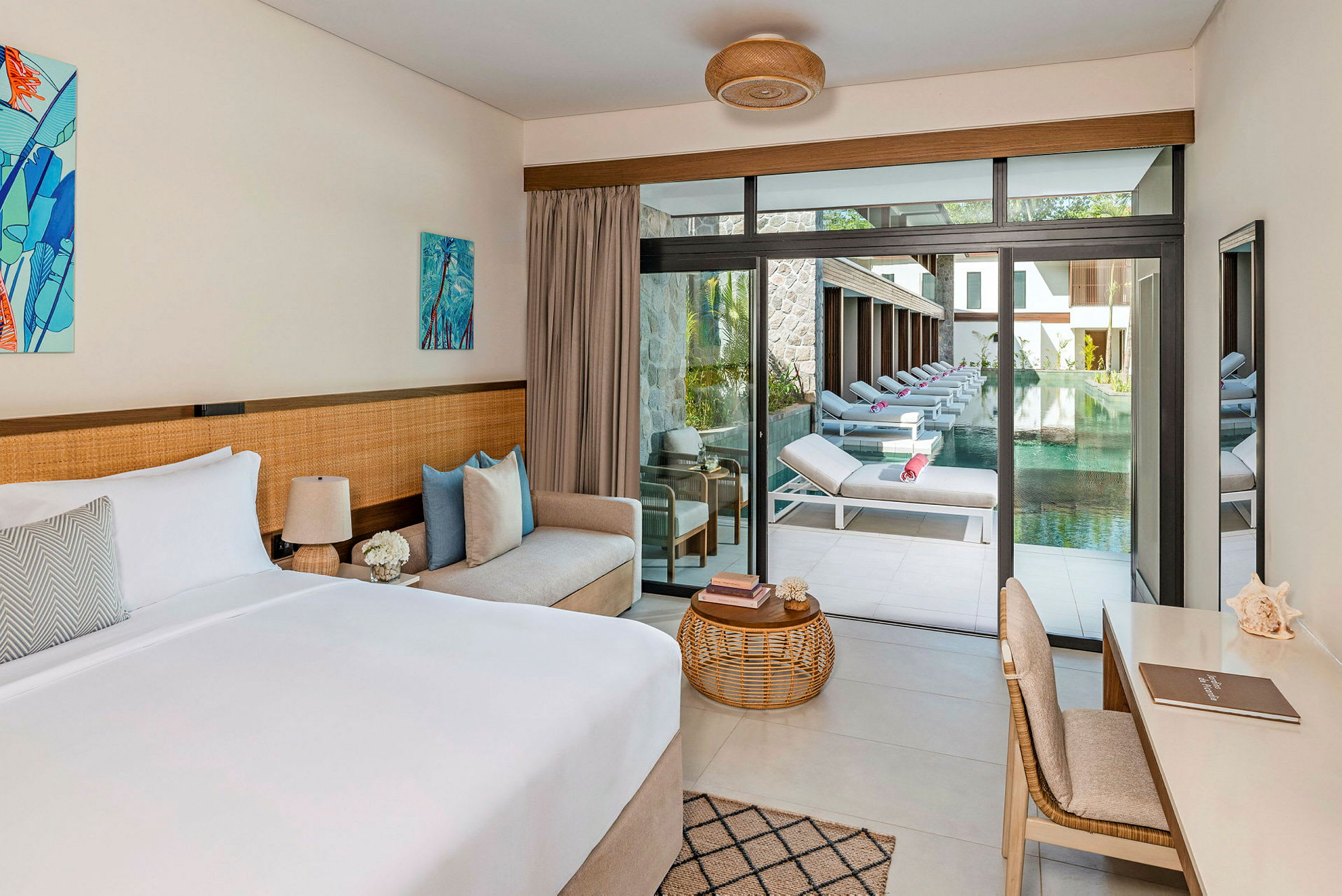 Ett modernt hotellrum med säng, soffa och skrivbord. Glasdörrar leder ut till en terrass med solstolar vid en pool. (AI-genererad text)