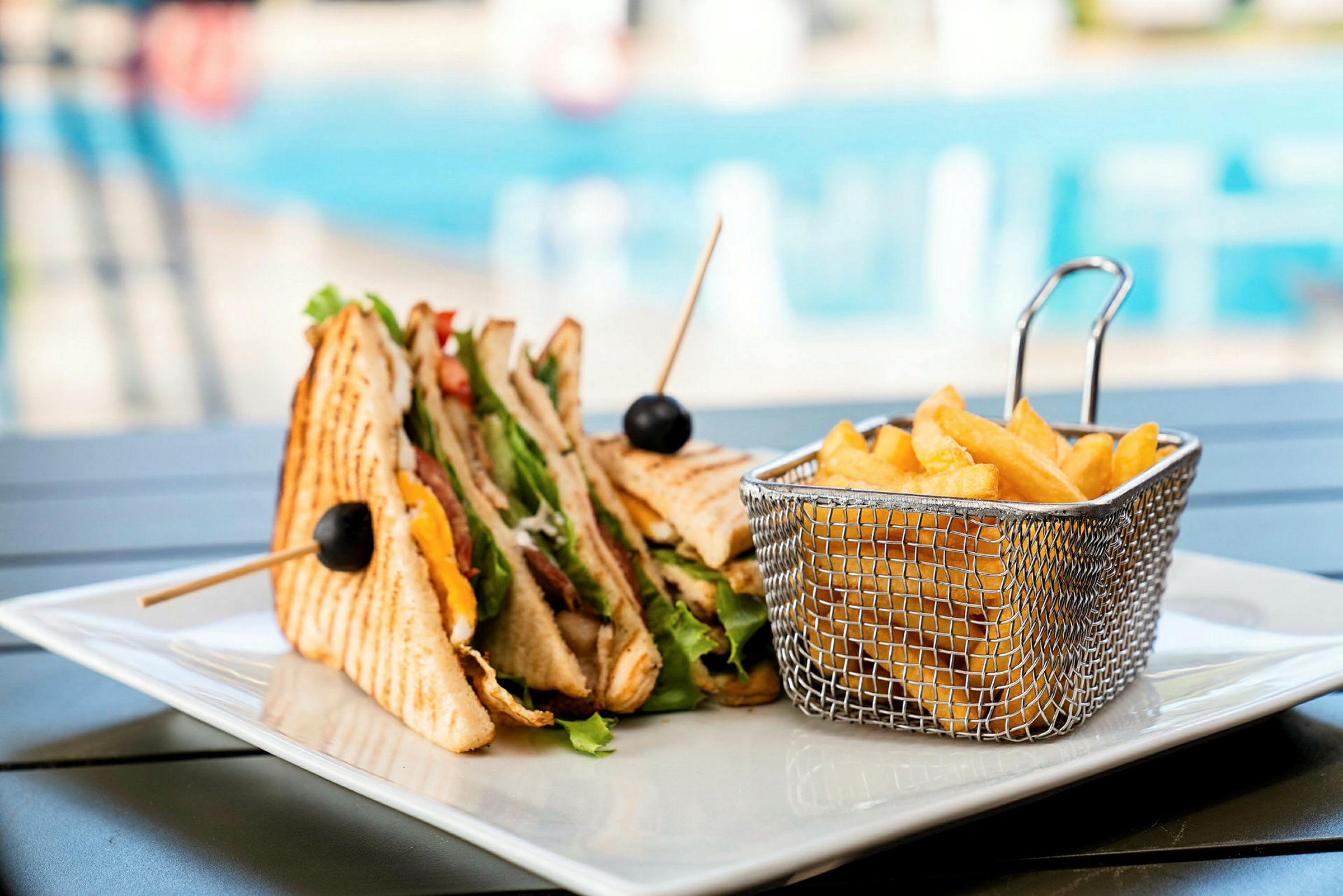 En tallrik med en trippel klubbsmörgås och en korg med pommes frites. I bakgrunden anas en swimmingpool. (AI-genererad text)
