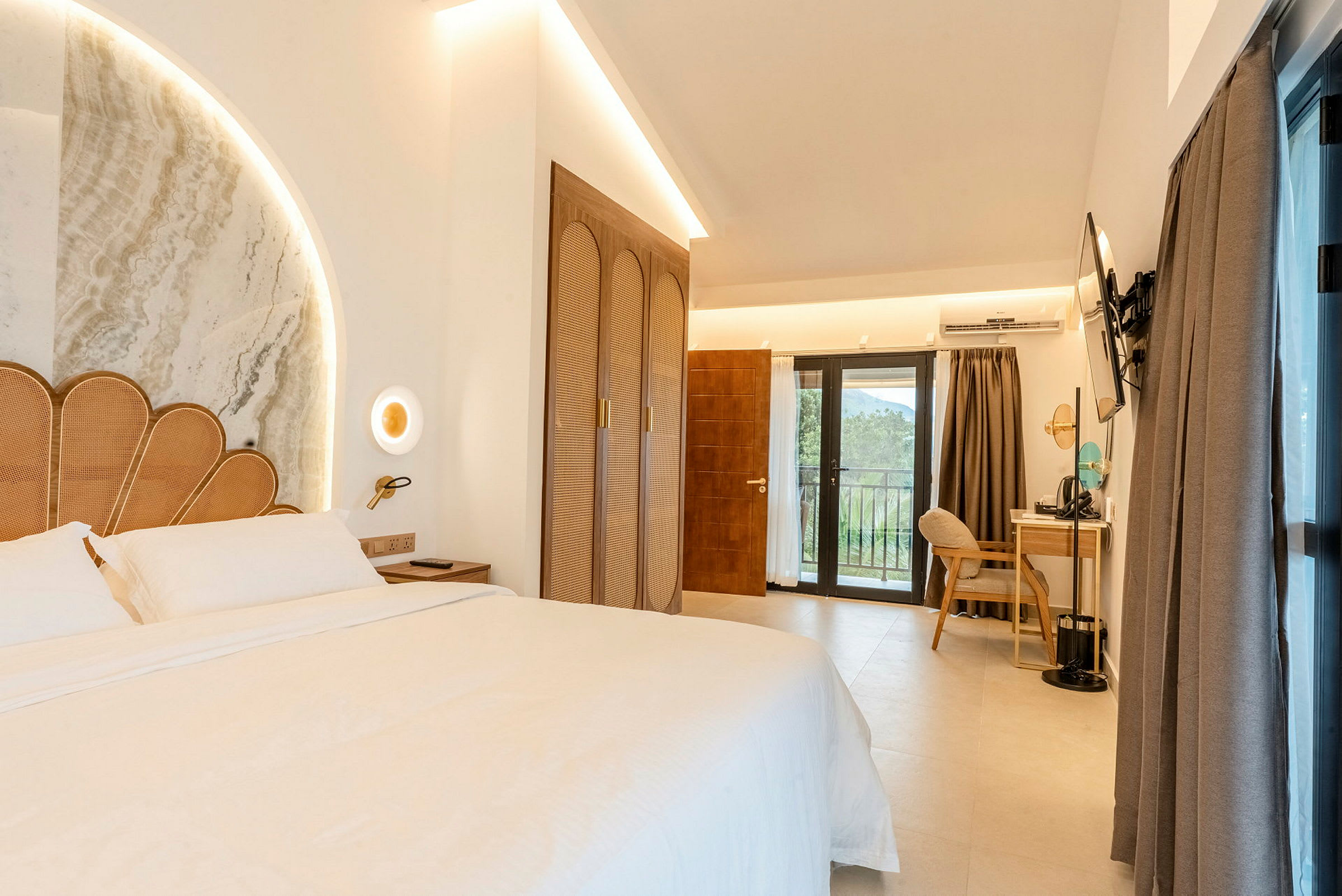 Ett hotellrum med en stor säng med dekorativ sänggavel och sängbord. Garderob, skrivbord med stol och TV, samt balkongdörr med utsikt. (AI-genererad text)