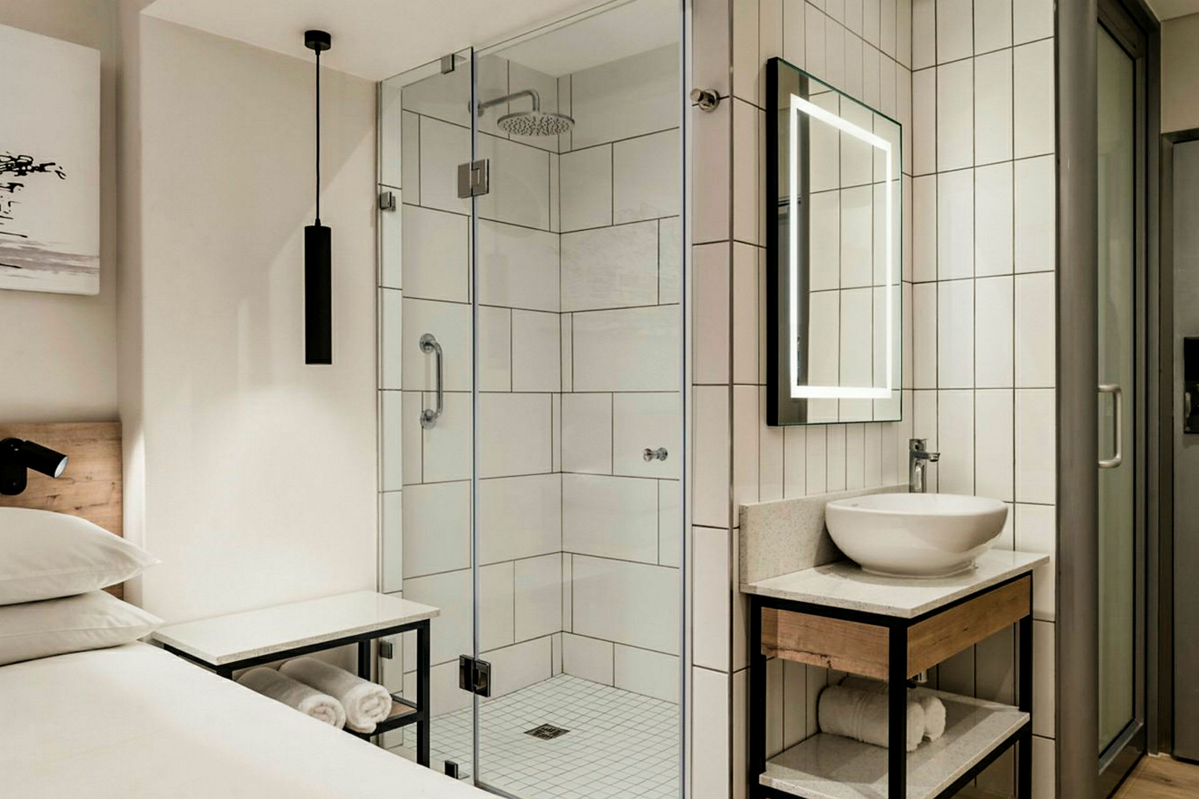 Ett hotellrum med en dusch med glasväggar, en spegel med belysning och en handfat på en modern möbel med handdukar under. (AI-genererad text)