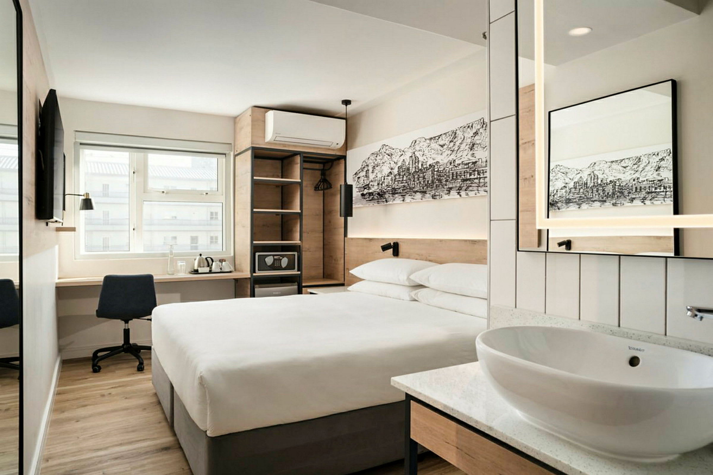 Ett modernt hotellrum med en dubbelsäng, ett skrivbord med stol, en väggmonterad TV, samt en öppen garderob och en liten tvättställ med spegel. (AI-genererad text)