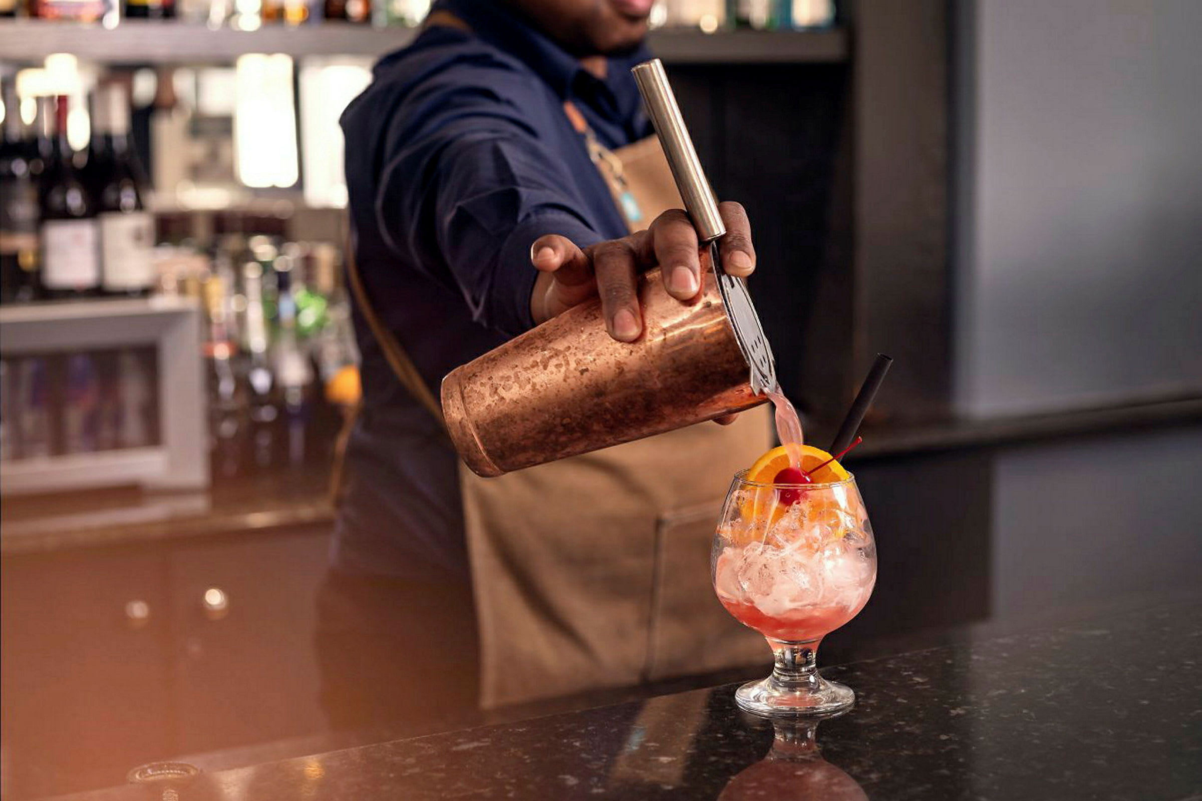 En bartender häller en rosa dryck från en shaker i ett glas med is och en dekoration av apelsinskiva och körsbär. (AI-genererad text)