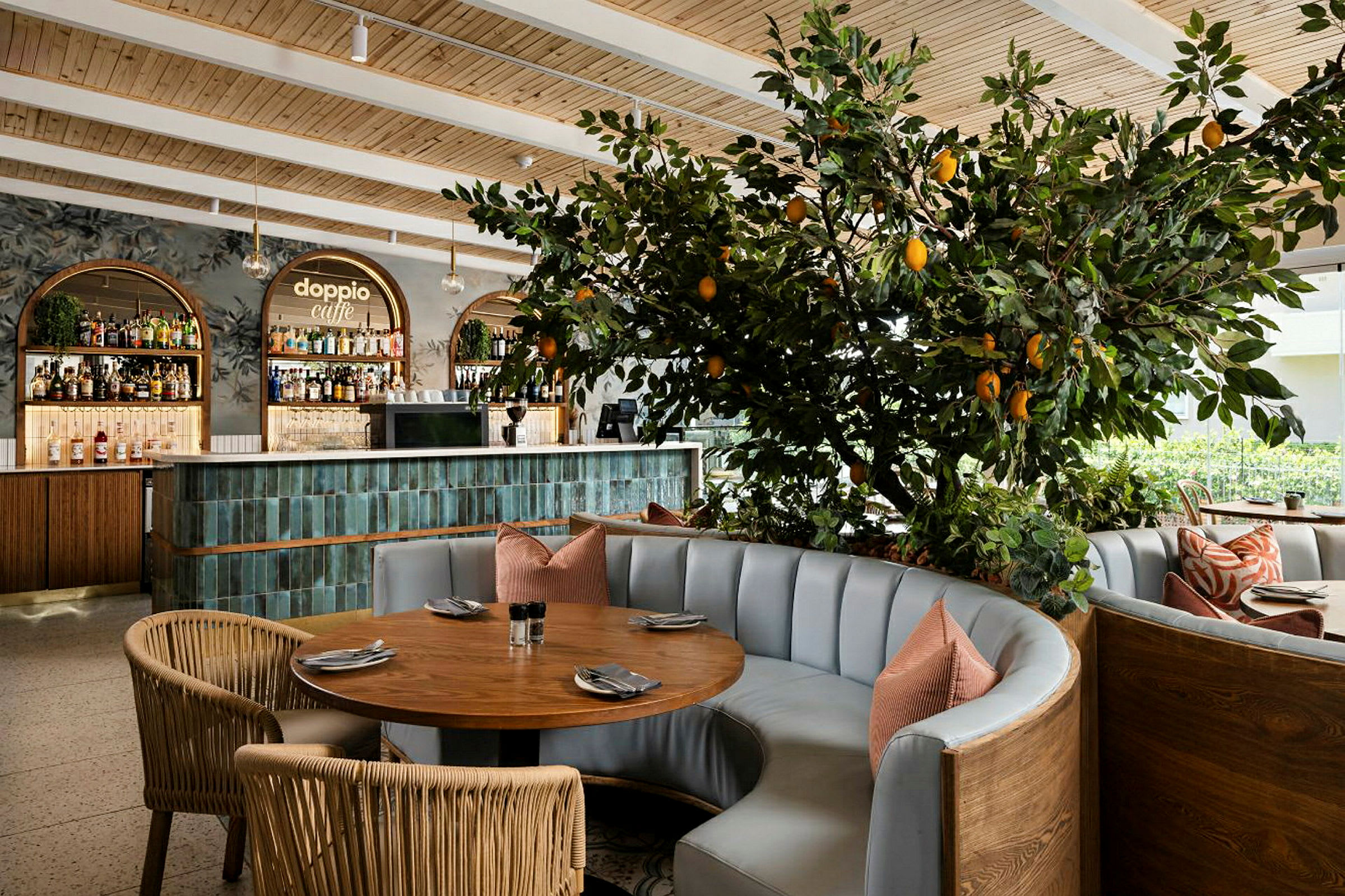 Interiör av en restaurang med ett runt bord, stoppade soffor och en dekorativt planterad citrusväxt med frukt. I bakgrunden syns en bardisk. (AI-genererad text)