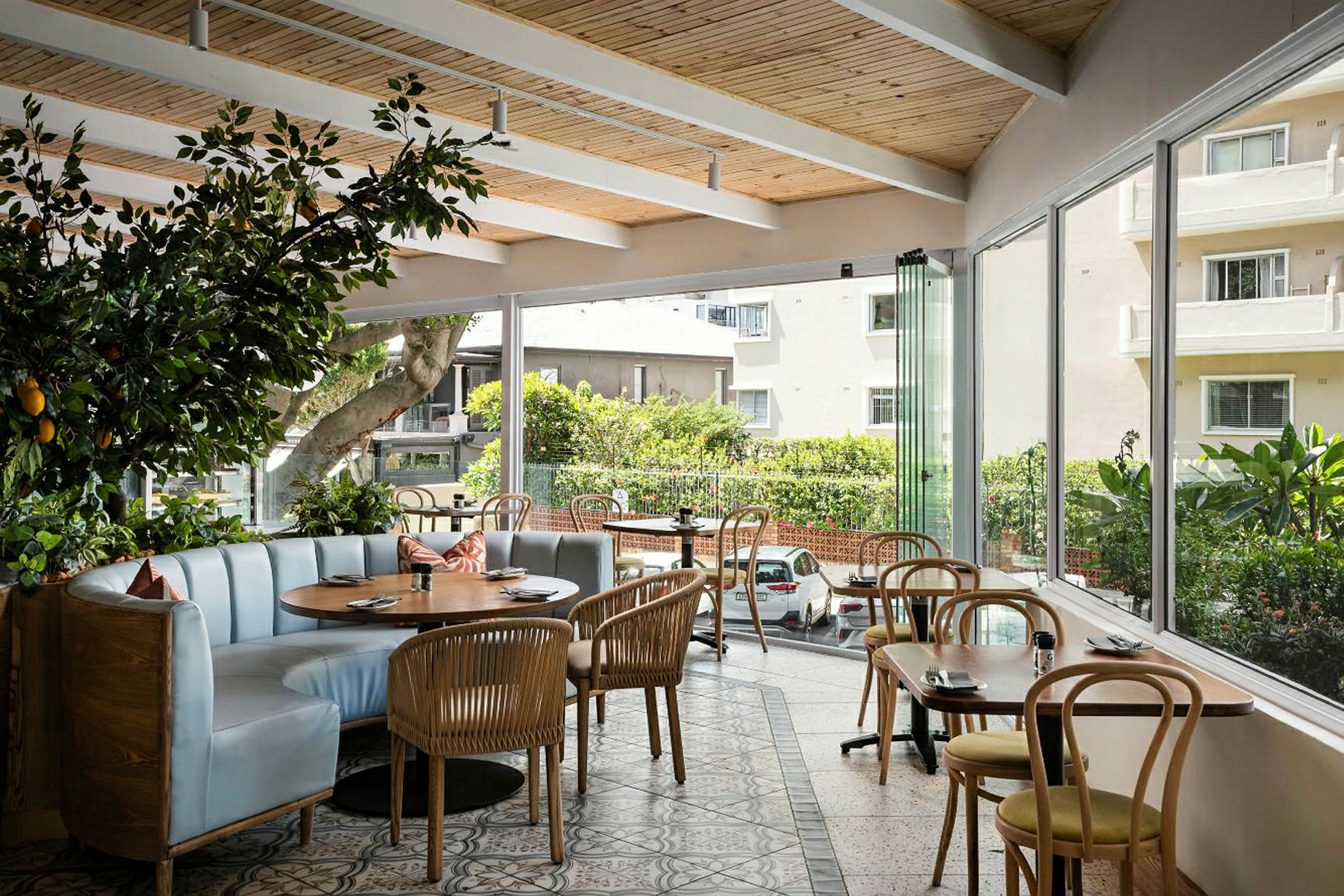 Ett café eller restaurang med stora fönster som släpper in mycket ljus. Lokalen har trädetaljer i taket, böjträstolar och ett långbord med stoppade sittplatser. (AI-genererad text)