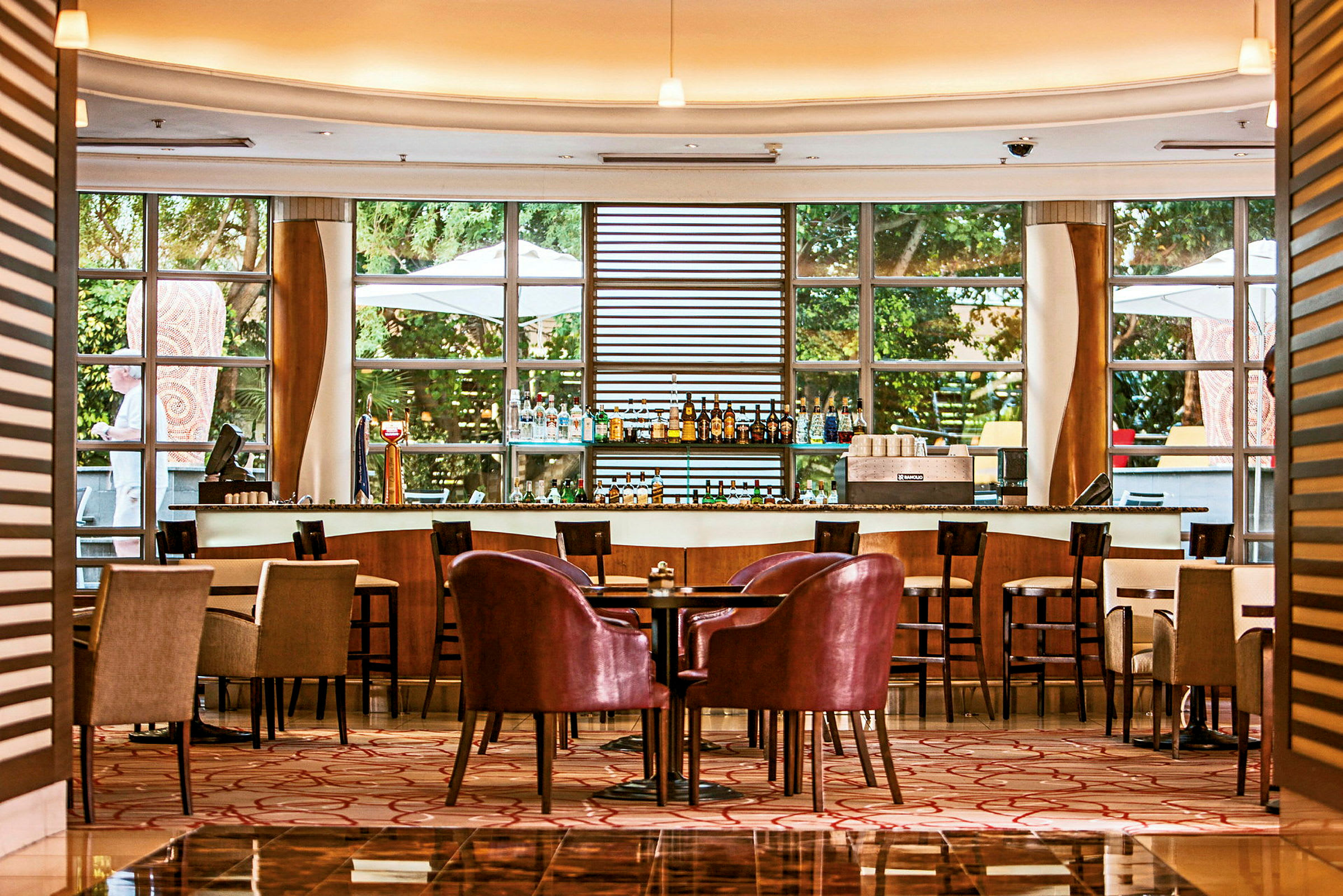 En elegant bar och sittområde med bord och stolar på ett hotell eller restaurang. Stora fönster släpper in mycket ljus utifrån. (AI-genererad text)