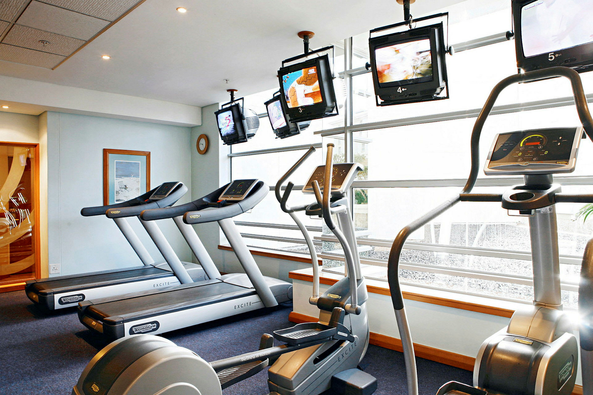 Bild av ett gym med träningsmaskiner som löpband och crosstrainers, samt tv-skärmar vid fönstren. (AI-genererad text)