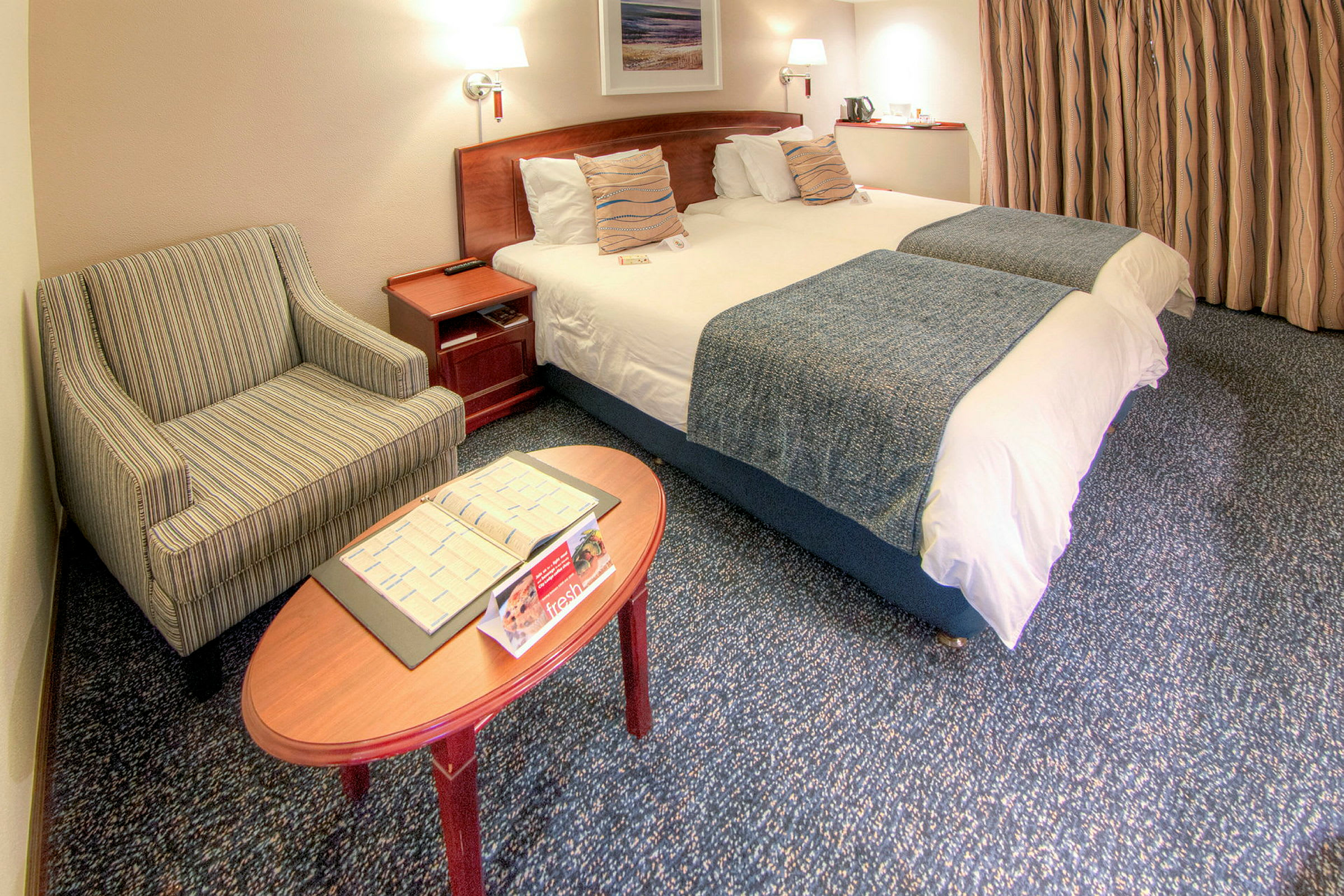 Ett hotellrum med två bäddade enkelsängar, en randig fåtölj och ett soffbord med broschyrer. (AI-genererad text)