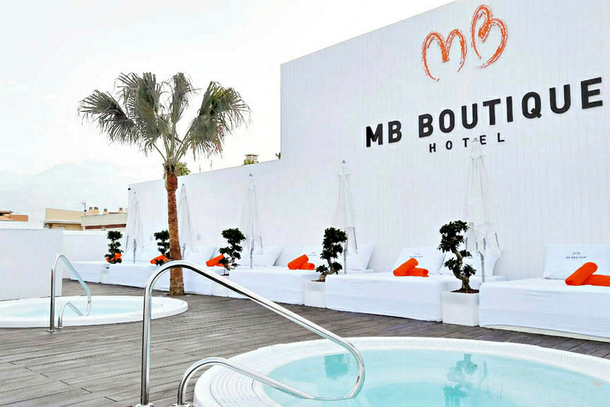 Ett hotellområde med vita solstolar och orange handdukar vid en pool, samt en palm och vita väggar. (AI-genererad text)