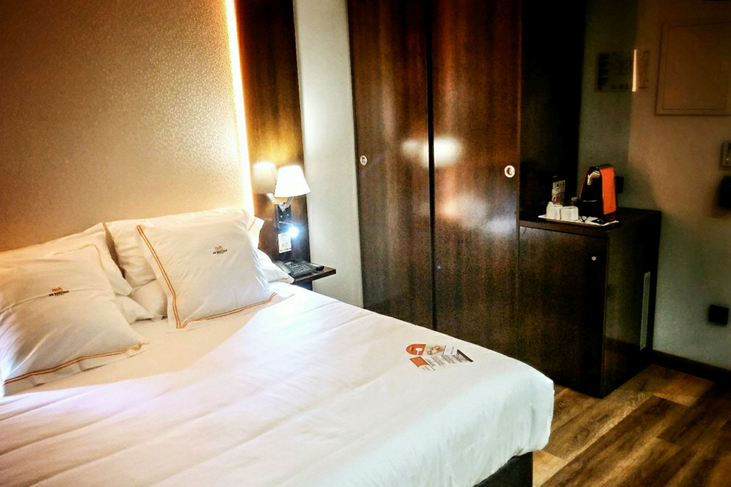 Ett hotellrum med en säng och upplyst nattlampa bredvid ett mörkt träskåp. (AI-genererad text)