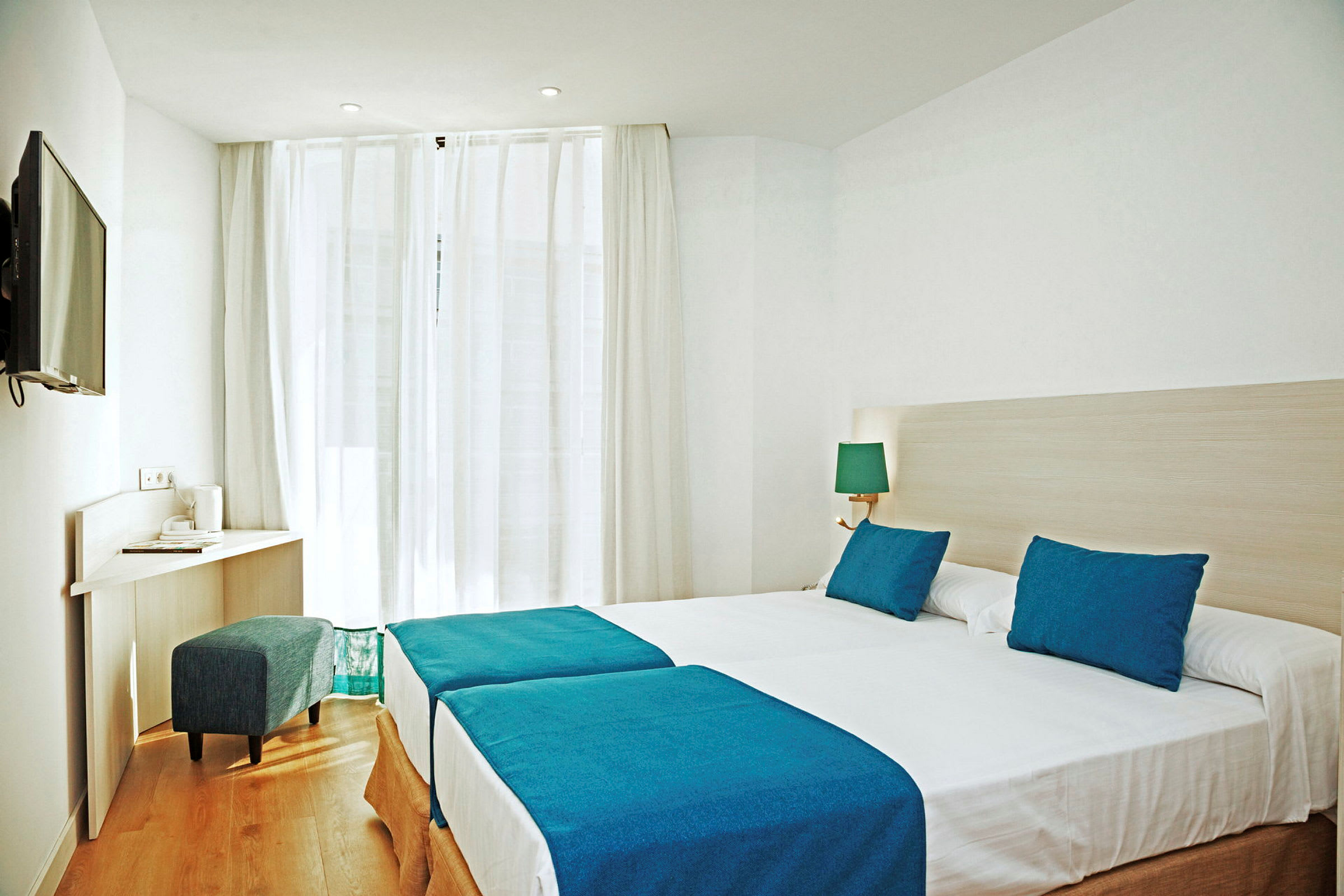 Ett hotellrum med två enkelsängar med blå sängöverkast och kuddar, en skrivbordshörna och en TV på väggen. (AI-genererad text)