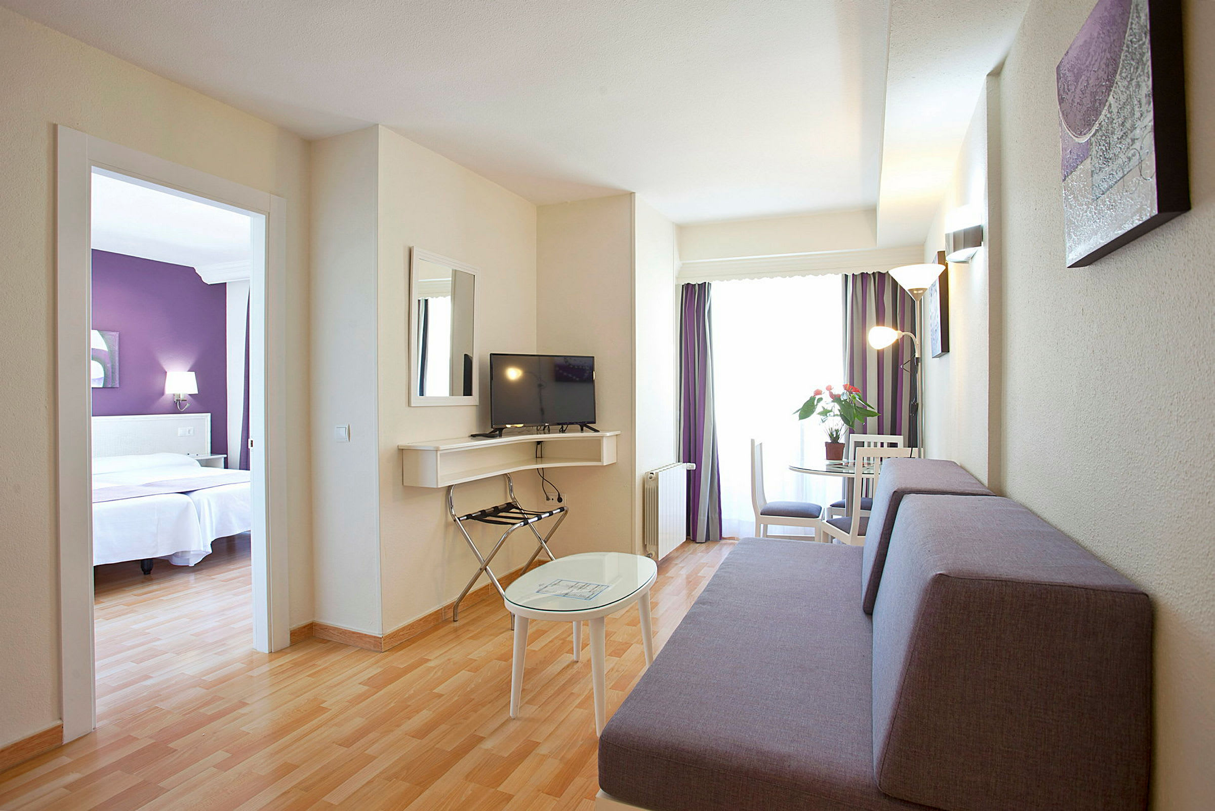 Hotellrum med vardagsrumsdel inklusive soffa, TV och bord, och separat sovrum synligt genom en dörröppning. (AI-genererad text)