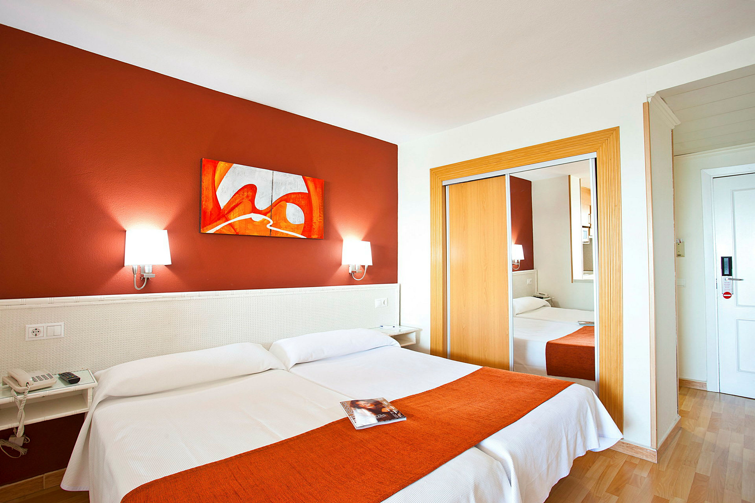 Ett hotellrum med två enkelsängar, en orange färgad löpare på sängen, en spegelgarderob, och väggdekor. (AI-genererad text)