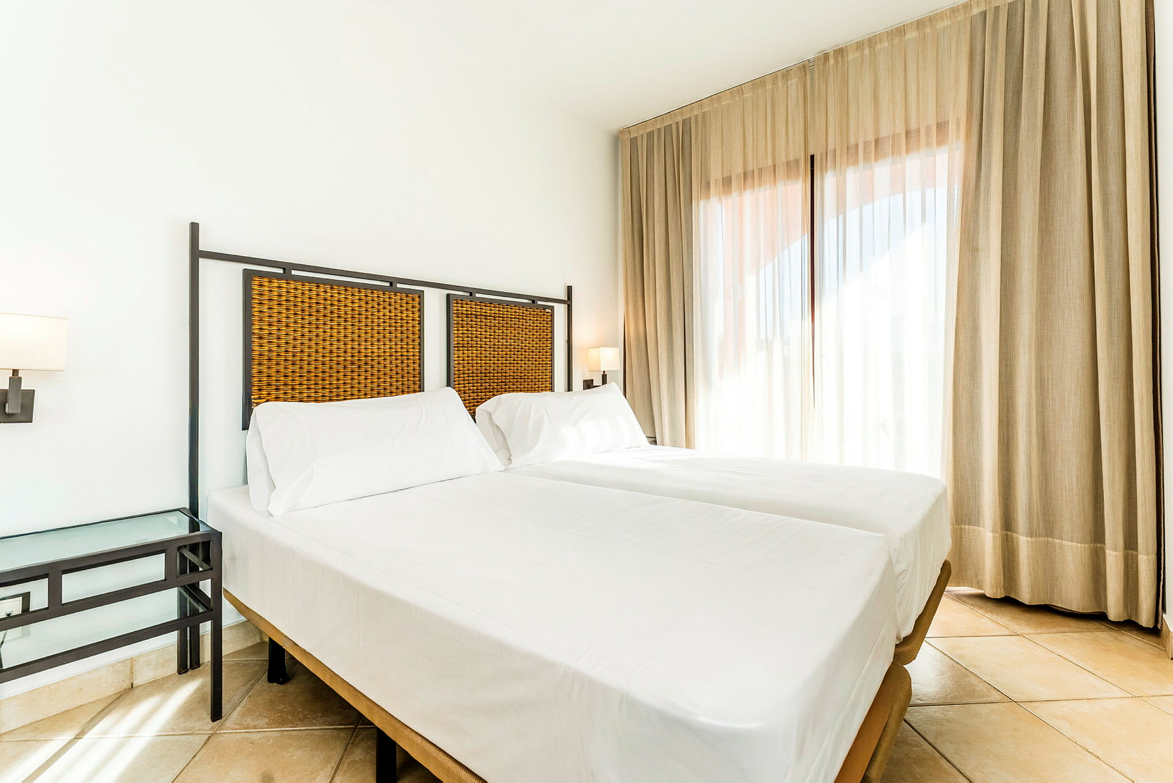 Ett ljust hotellrum med två enkelsängar, vita lakan och en flätad sänggavel. Rummet har ett sidobord och stora fönster med gardiner. (AI-genererad text)