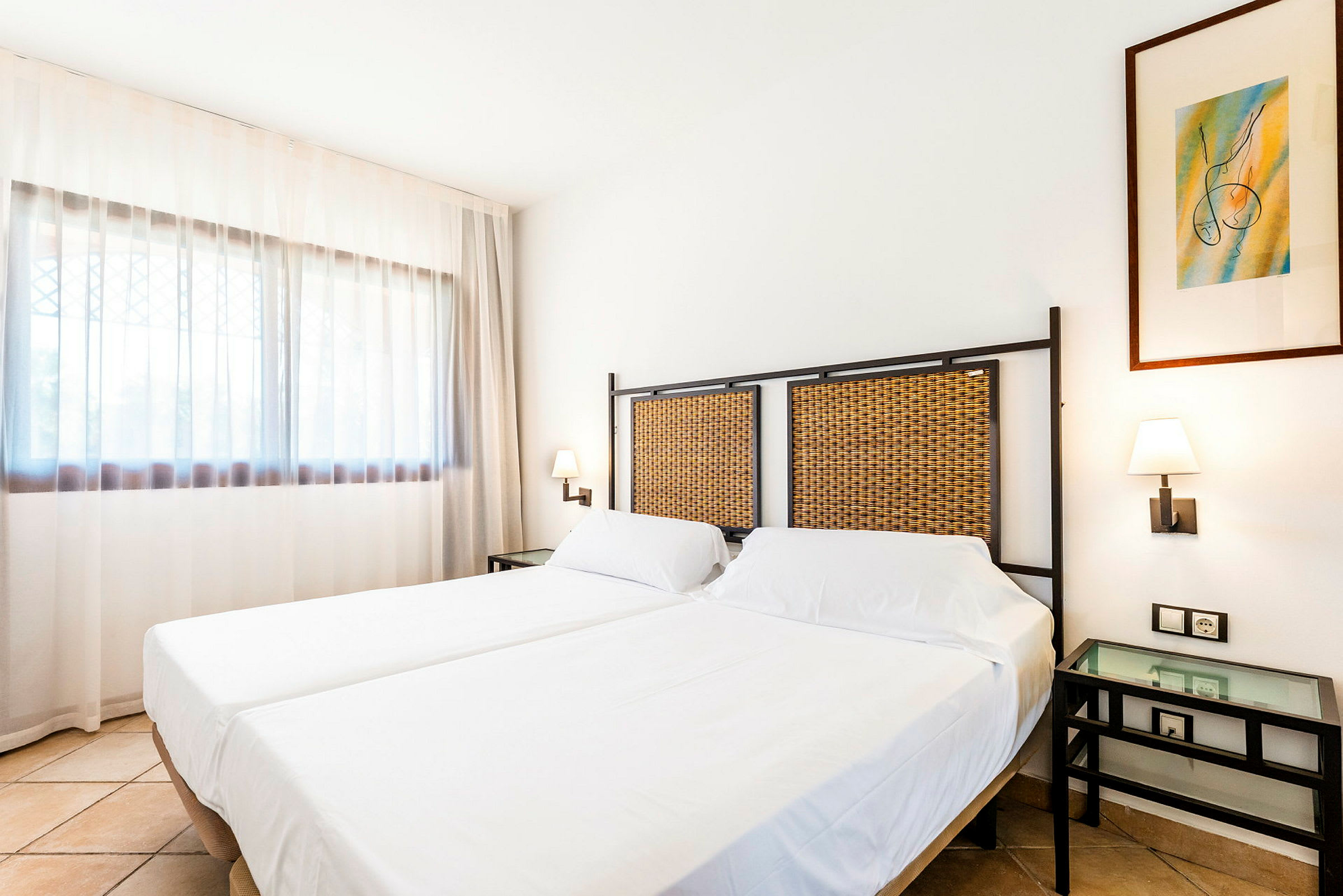 Ett hotellrum med två enkelsängar bredvid varandra, vita sängkläder, sänggavlar av flätat material och en nattlampa på väggen samt ett fönster med vita gardiner. (AI-genererad text)