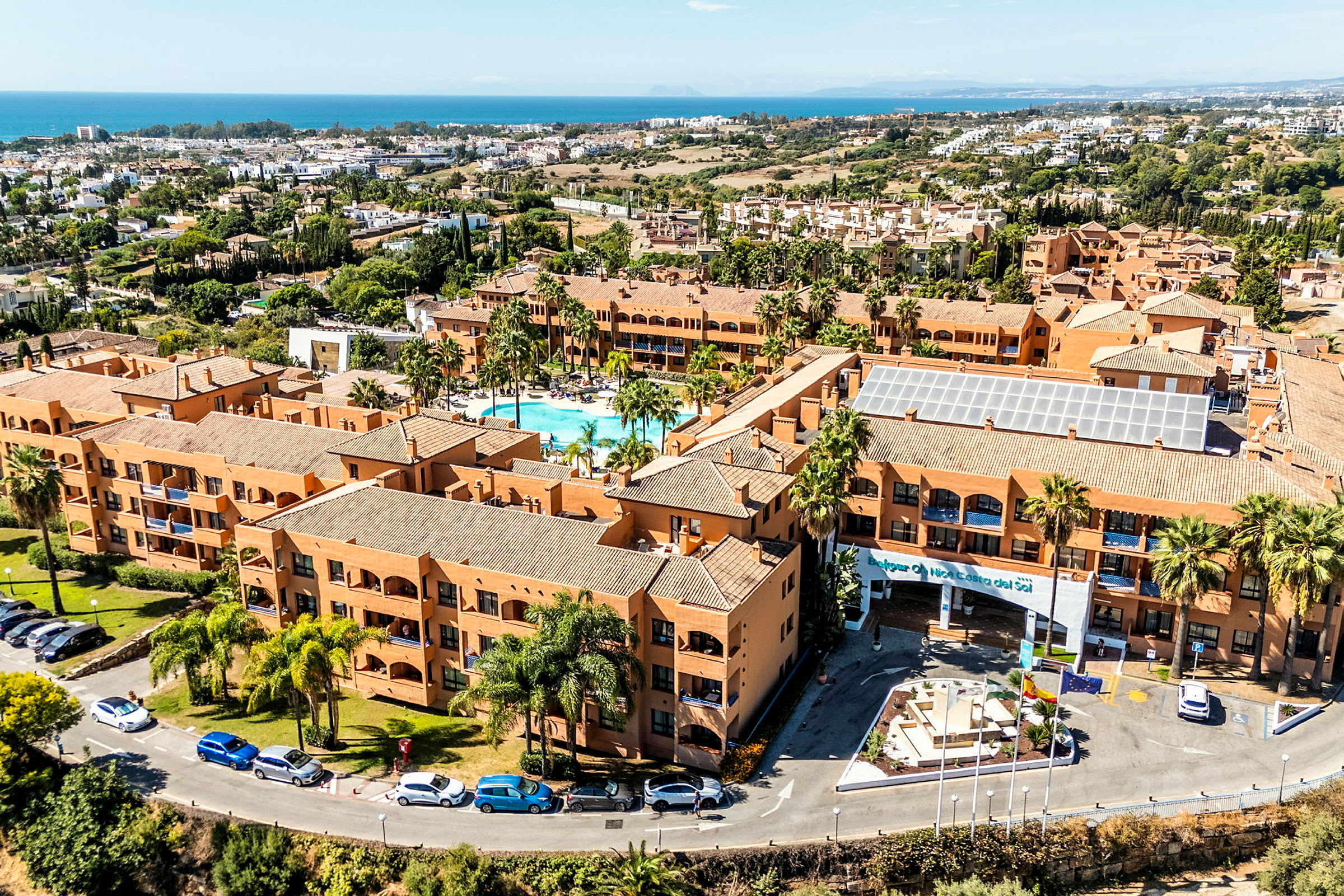 Ett semesterresort med terrakottafärgade byggnader, en pool i centrum och omgivande grönska. Havet syns i bakgrunden. (AI-genererad text)