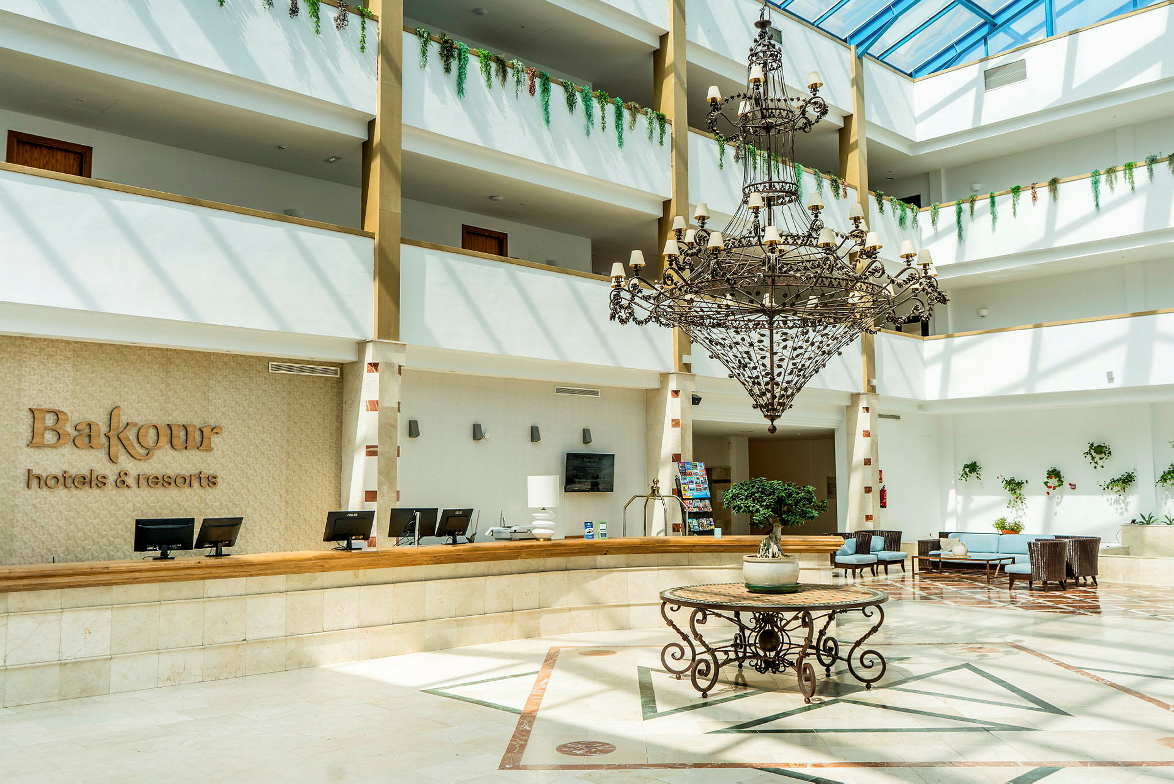 Bild av en hotellobby med receptionsdisk, en stor ljuskrona och en dekorativ växt på ett bord (AI-genererad text)
