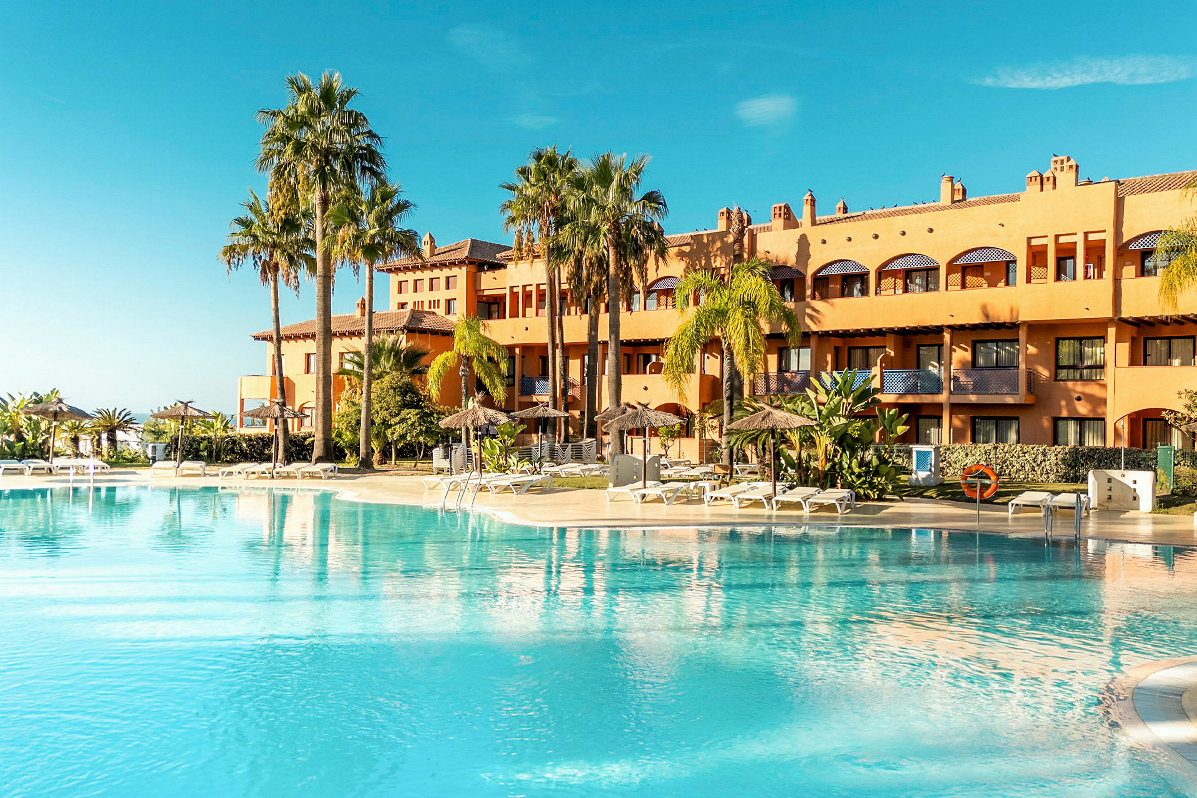 En stor utomhuspool framför ett hotell omgivet av palmer och solsängar. (AI-genererad text)