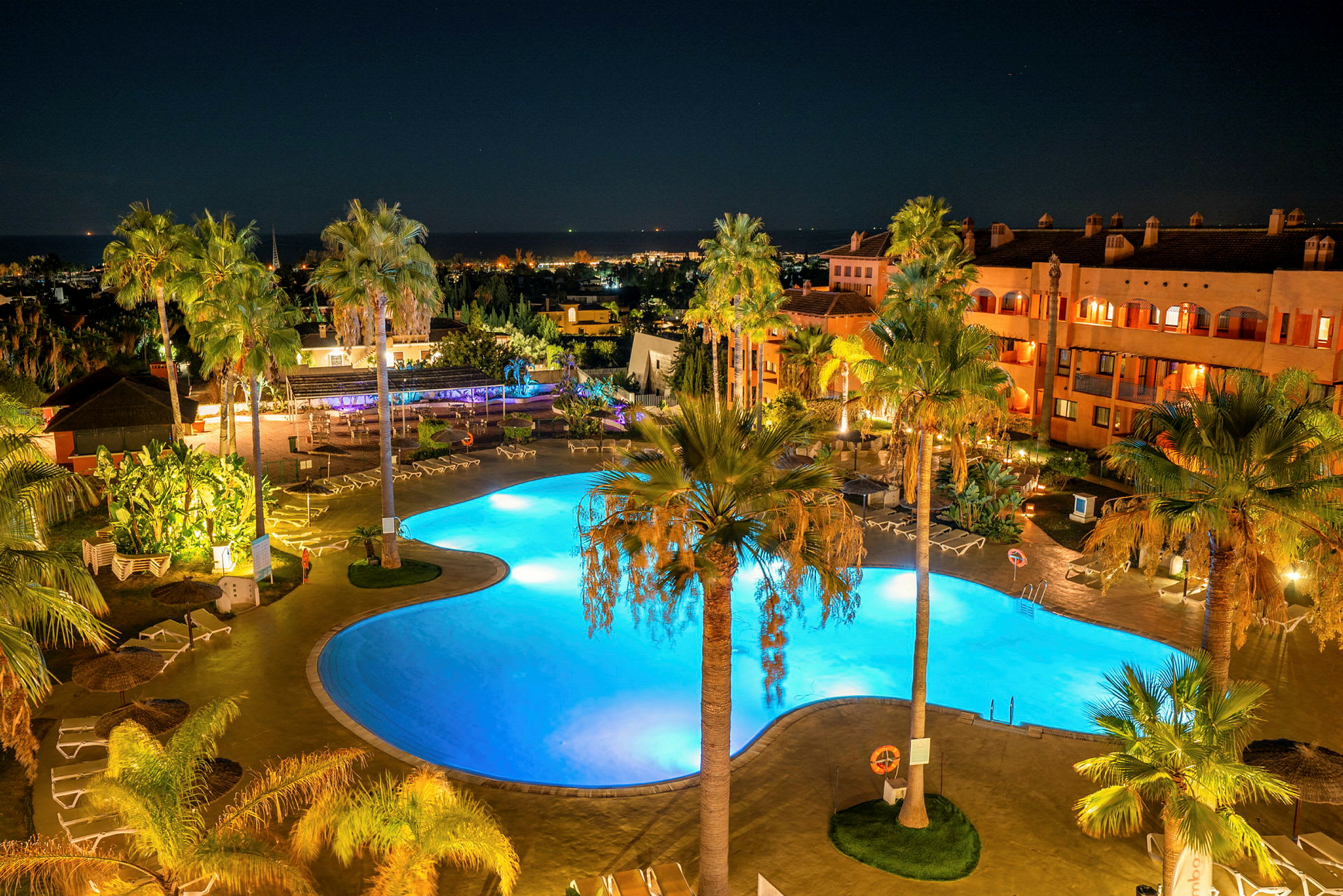 En belyst pool på en resort omgiven av palmer under natthimlen. (AI-genererad text)