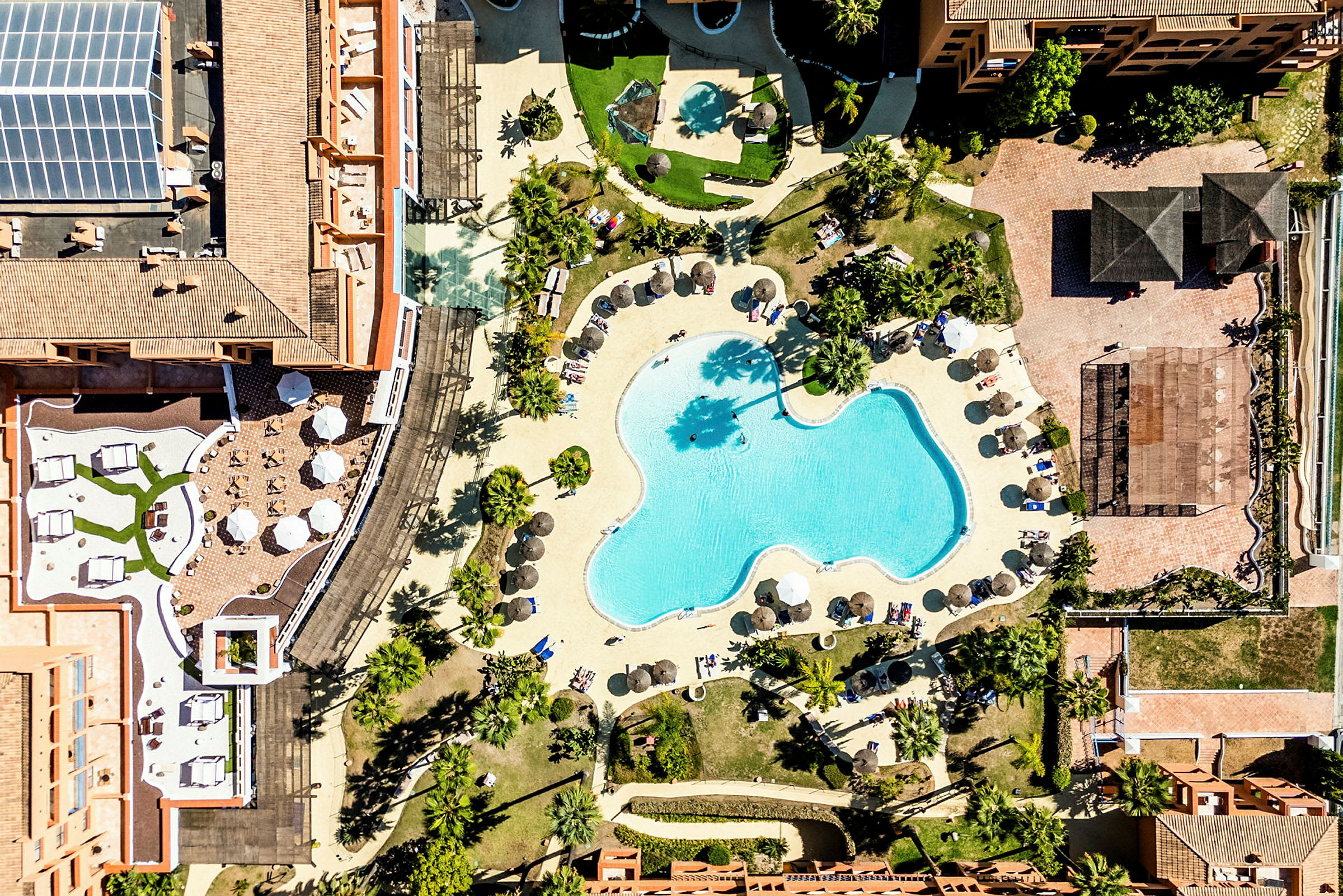Flygvy av ett hotellområde med en stor pool och omgivande solstolar och parasoll. (AI-genererad text)