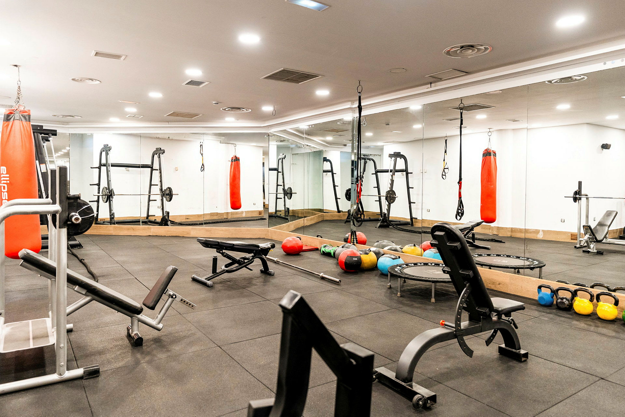 Ett gym med träningsutrustning inklusive bänkar, skivstänger, vikter, medicinbollar, kettlebells och boxsäckar. (AI-genererad text)