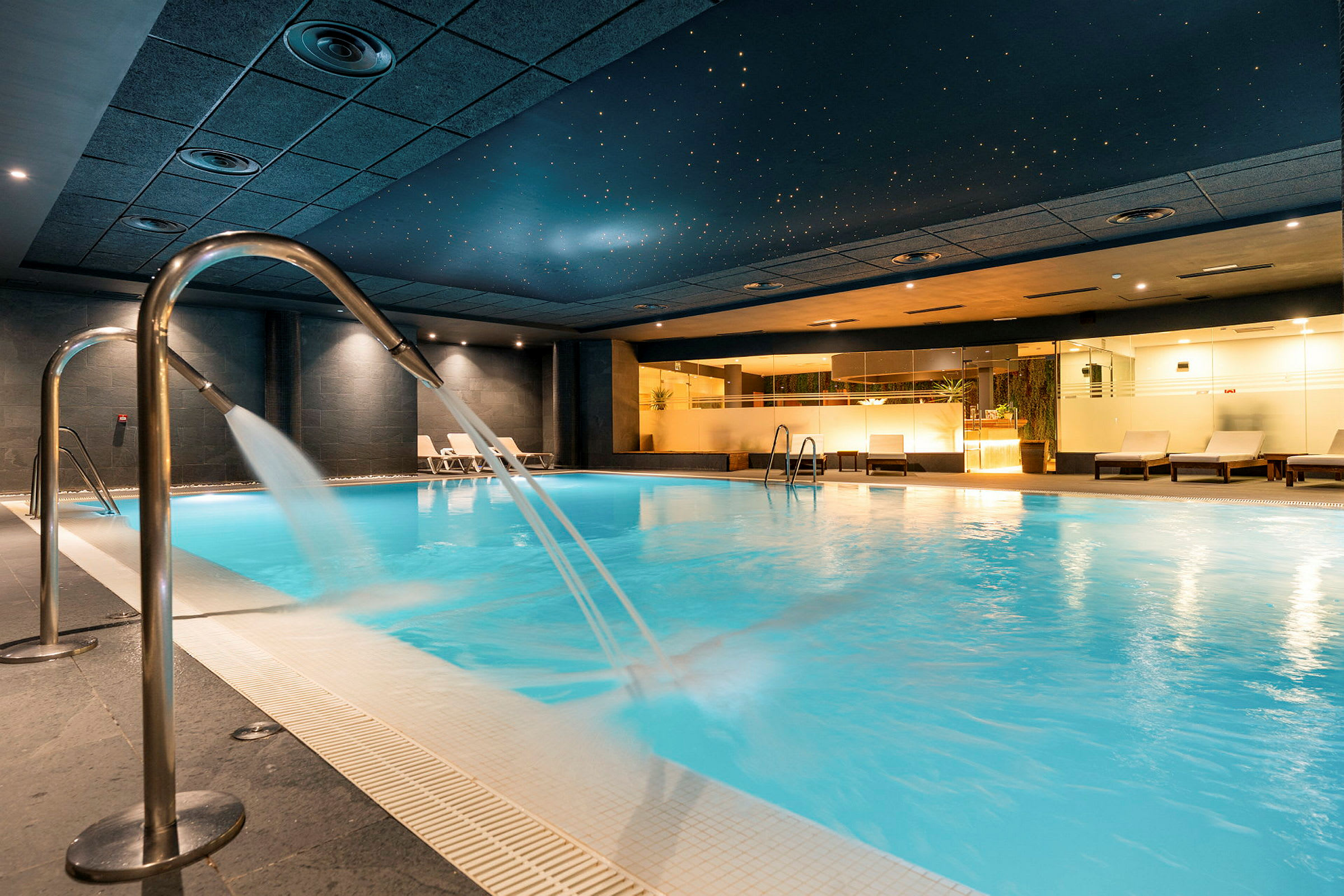 Inomhuspool med bäckar av vatten som flödar från metallstålar och avslappningsstolar vid poolkanten. (AI-genererad text)