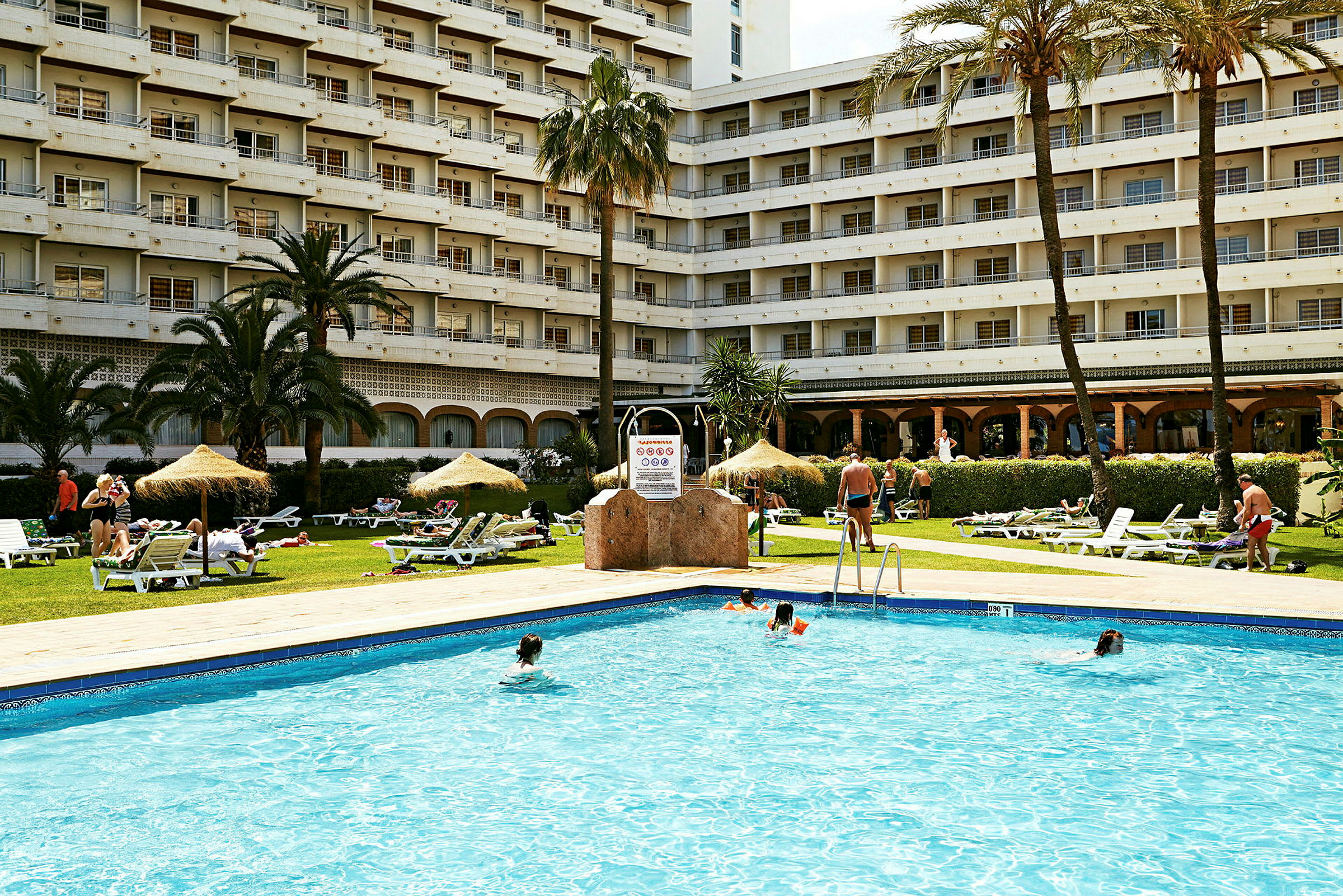 Utomhuspool med gräsmatta, parasoller, solstolar och hotellbyggnad i bakgrunden. (AI-genererad text)
