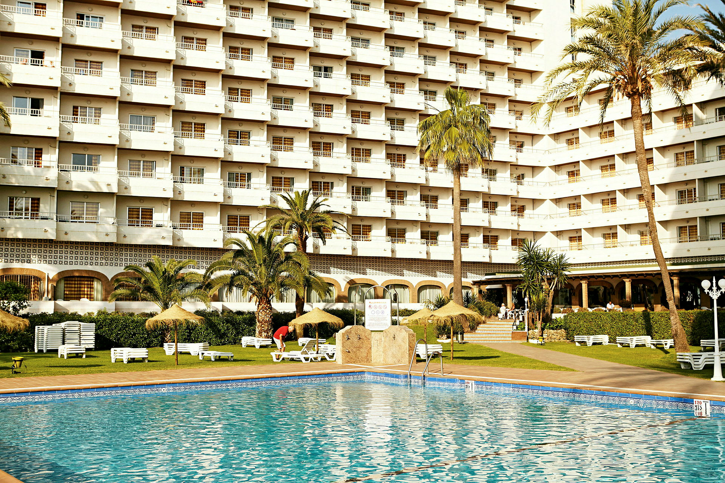 Hotellbyggnad med balkonger, palmträd och en pool i förgrunden. (AI-genererad text)