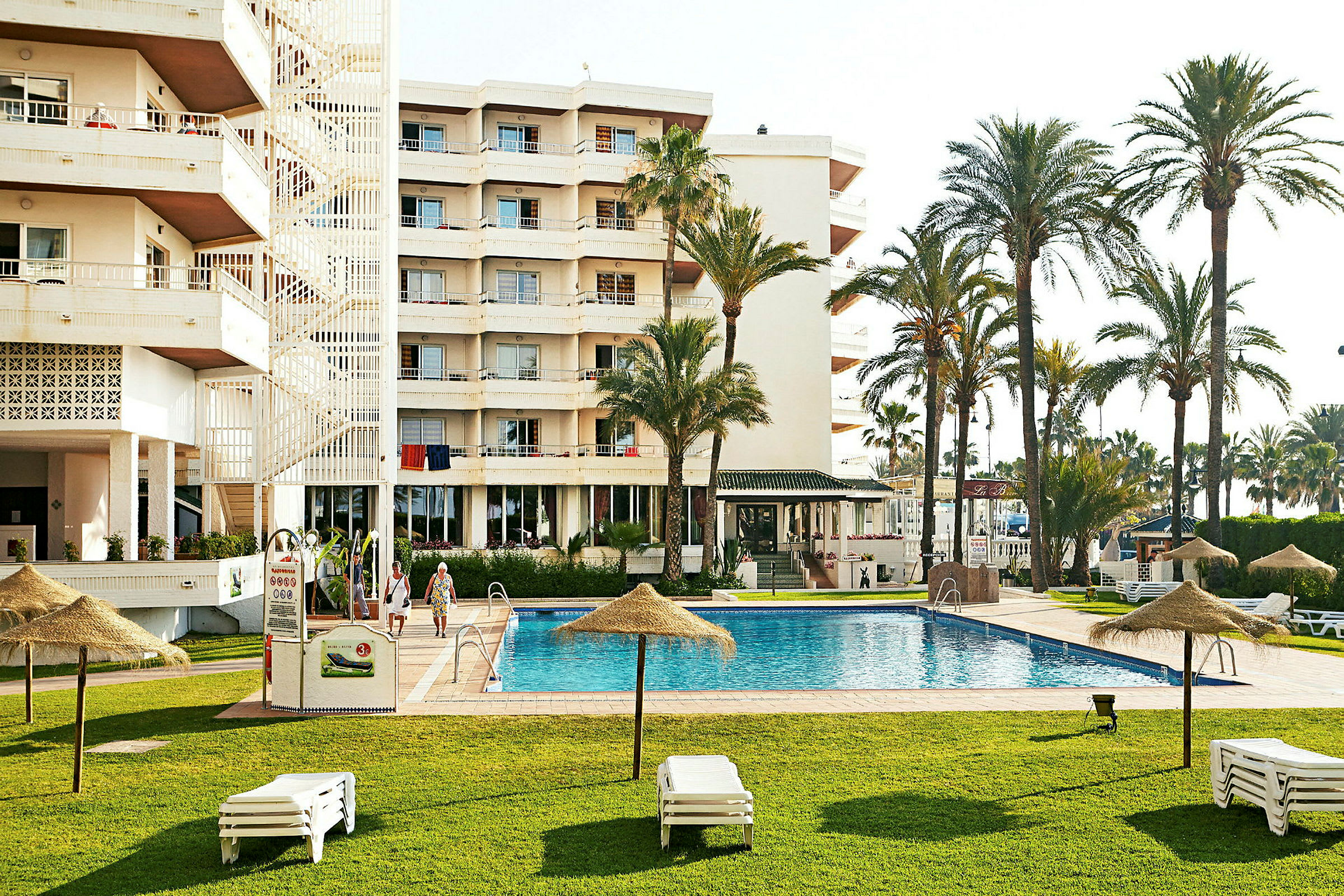 Hotellbyggnad med pool, solstolar och palmer i förgrunden. (AI-genererad text)