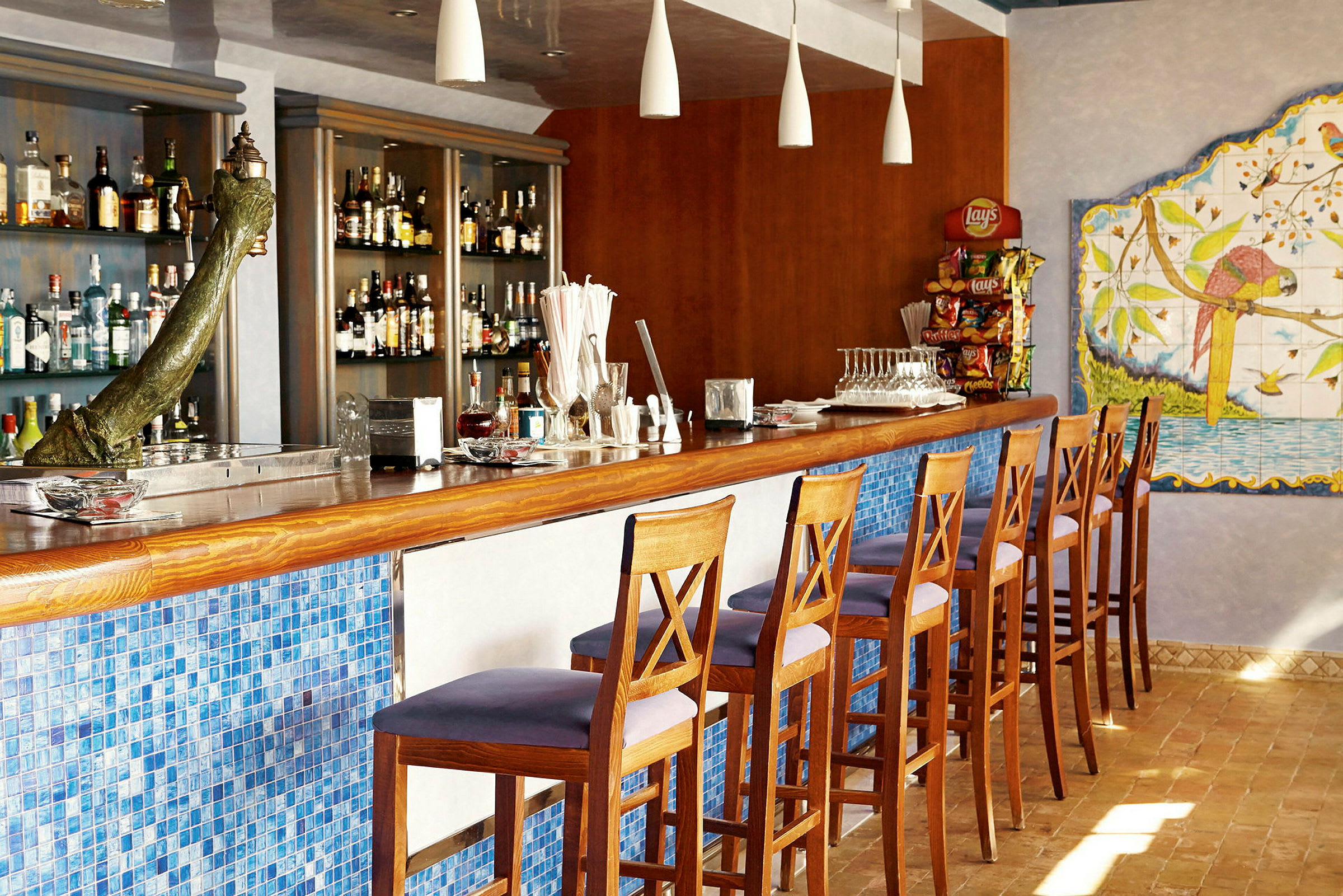 En bar med höga trästolar, en mosaikfront i blå färg, en imponerande samling av spritflaskor och en väggdekoration med en papegoja. (AI-genererad text)