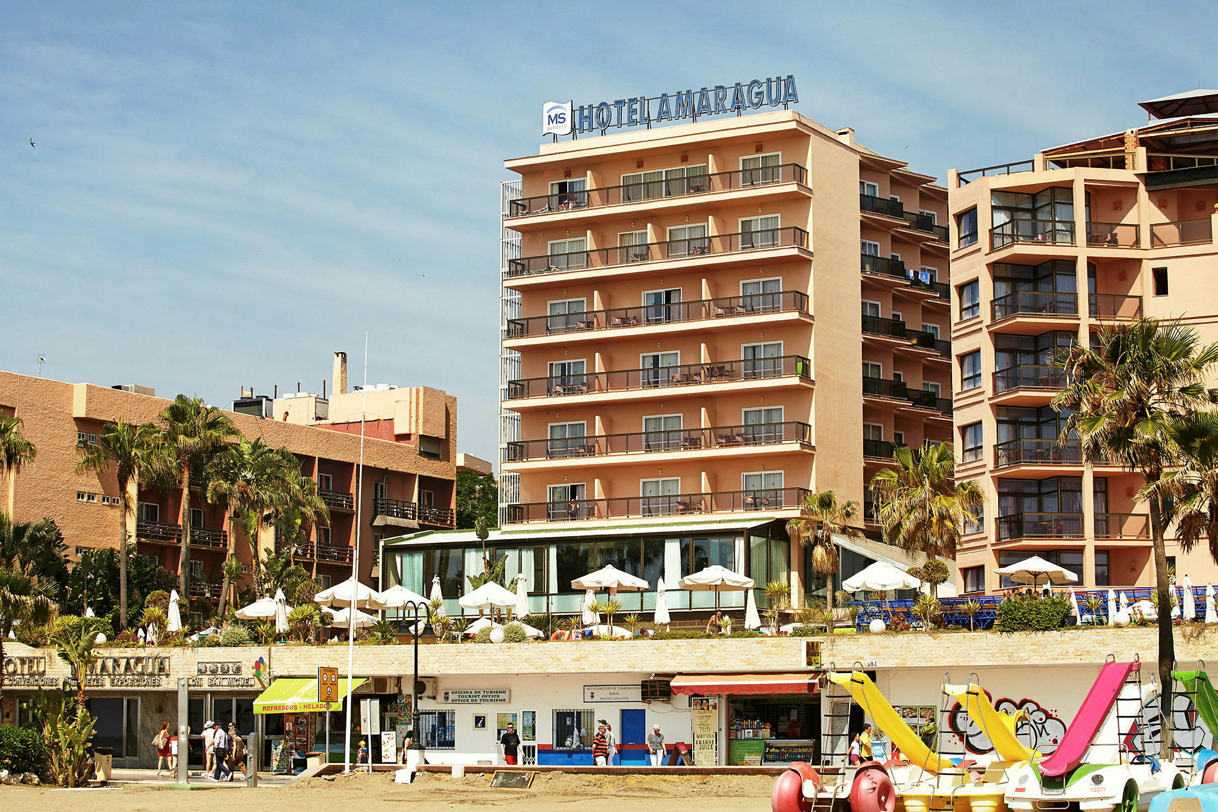 Ett hotell beläget vid stranden med parasoller och utsikt över havet. (AI-genererad text)