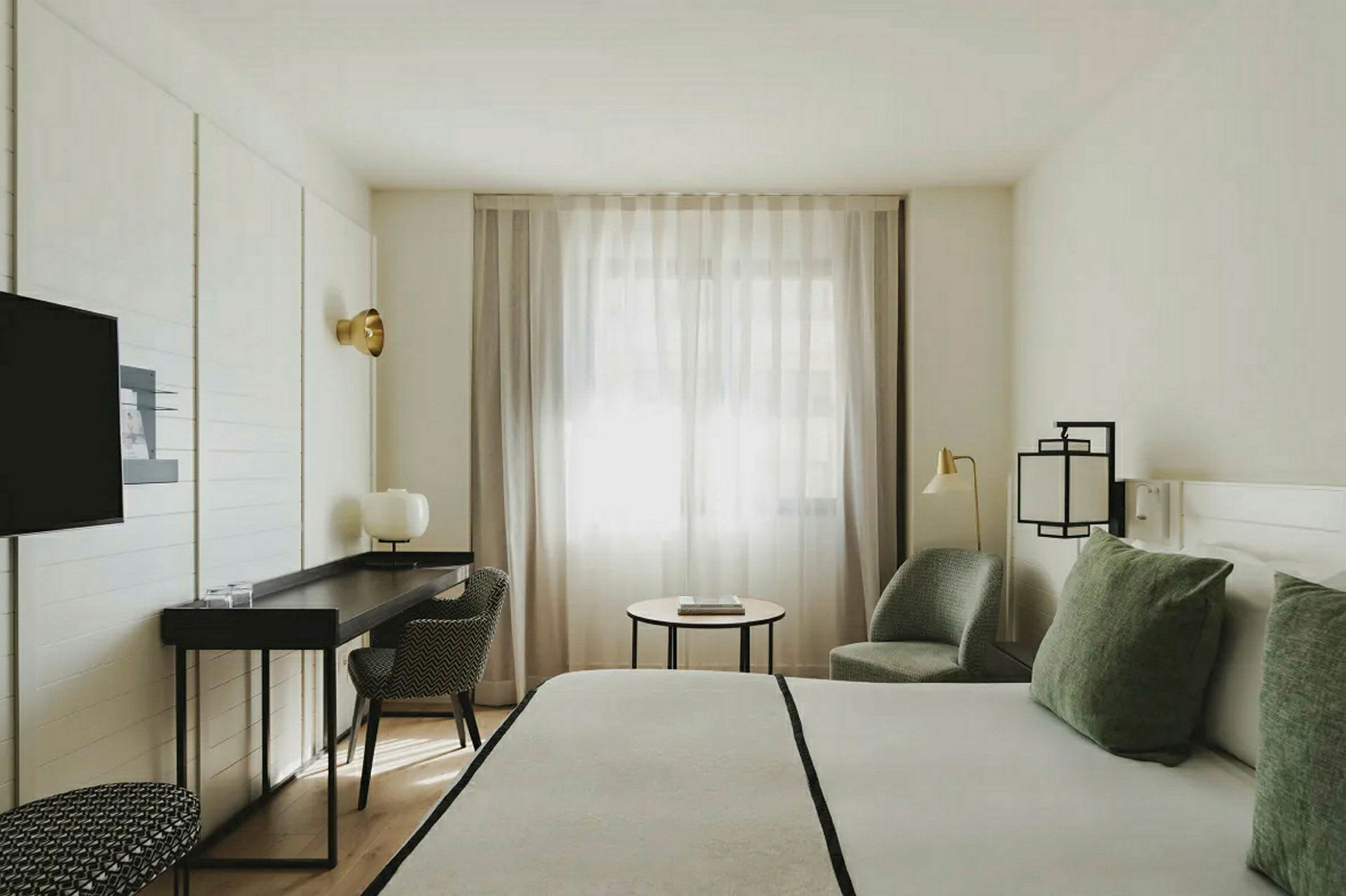 Ett ljust hotellrum med en stor säng, skrivbord, stol och fåtölj. Fönstret har tunna gardiner. (AI-genererad text)