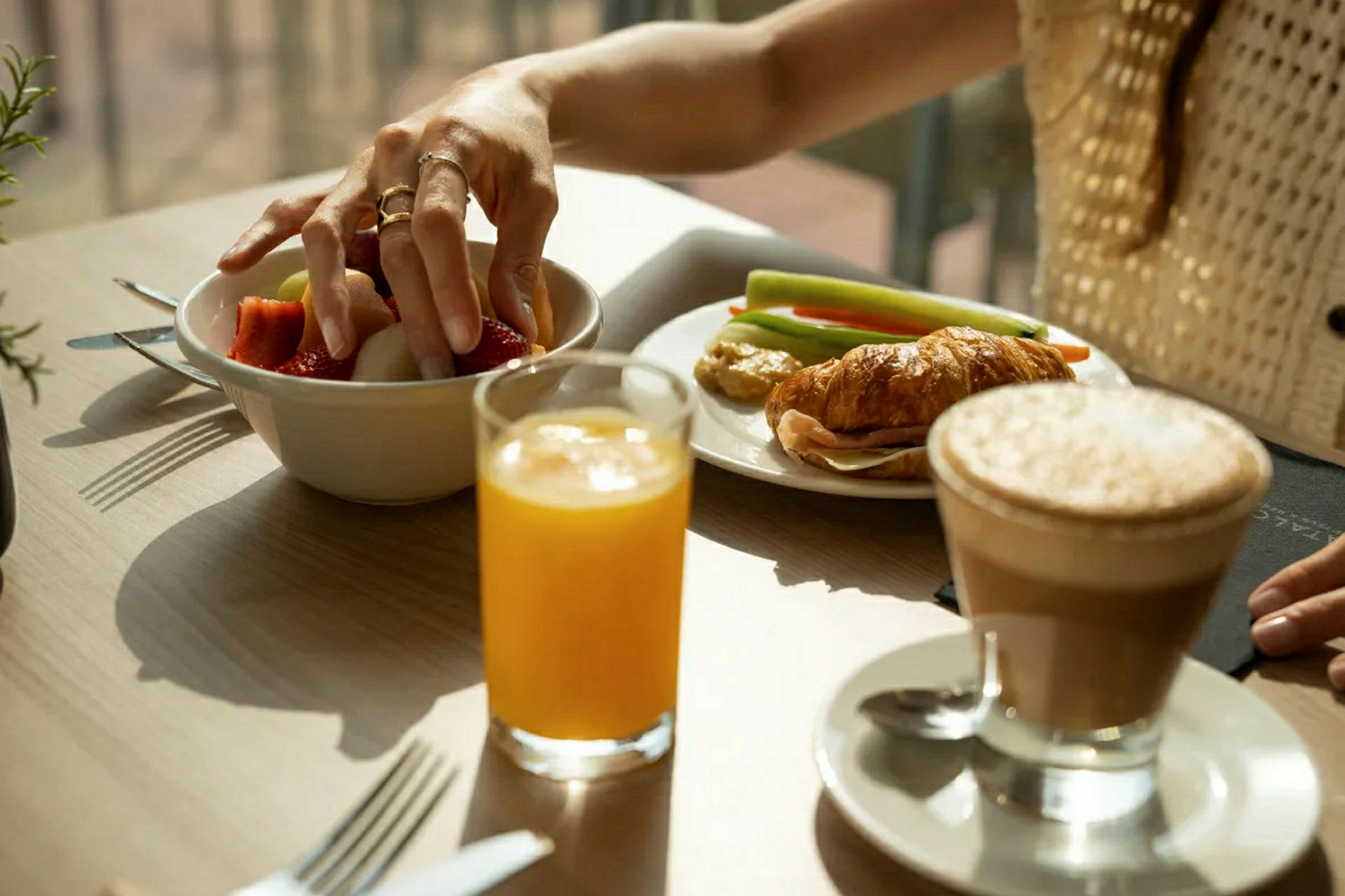Person tar frukt från en skål på ett bord med croissant, grönsaker, juice och kaffe. (AI-genererad text)