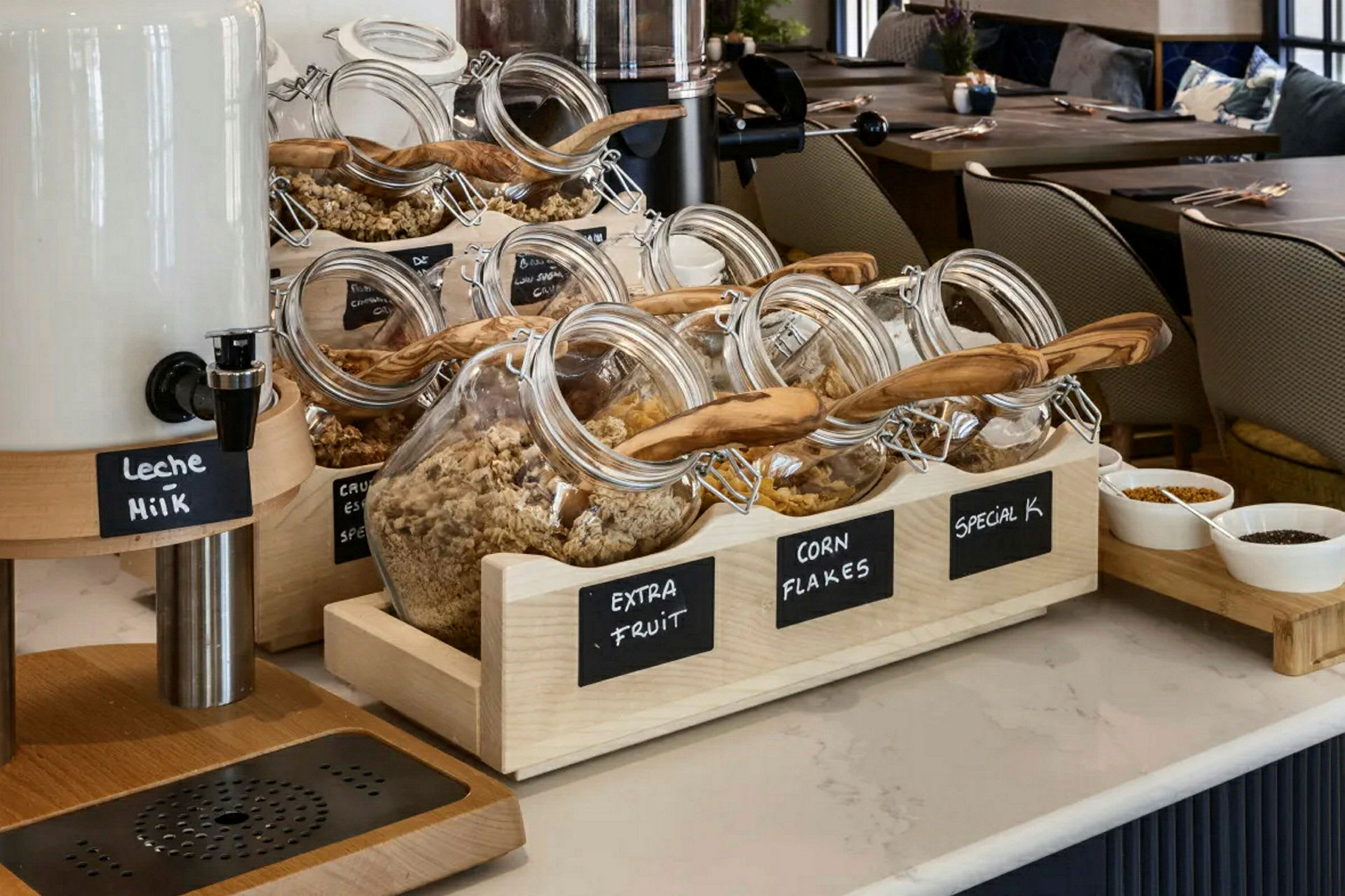 Glasburkar fyllda med olika sorters flingor och müsliblandningar på ett hotellbarbord med mjölkflaska och skålar med topping. (AI-genererad text)