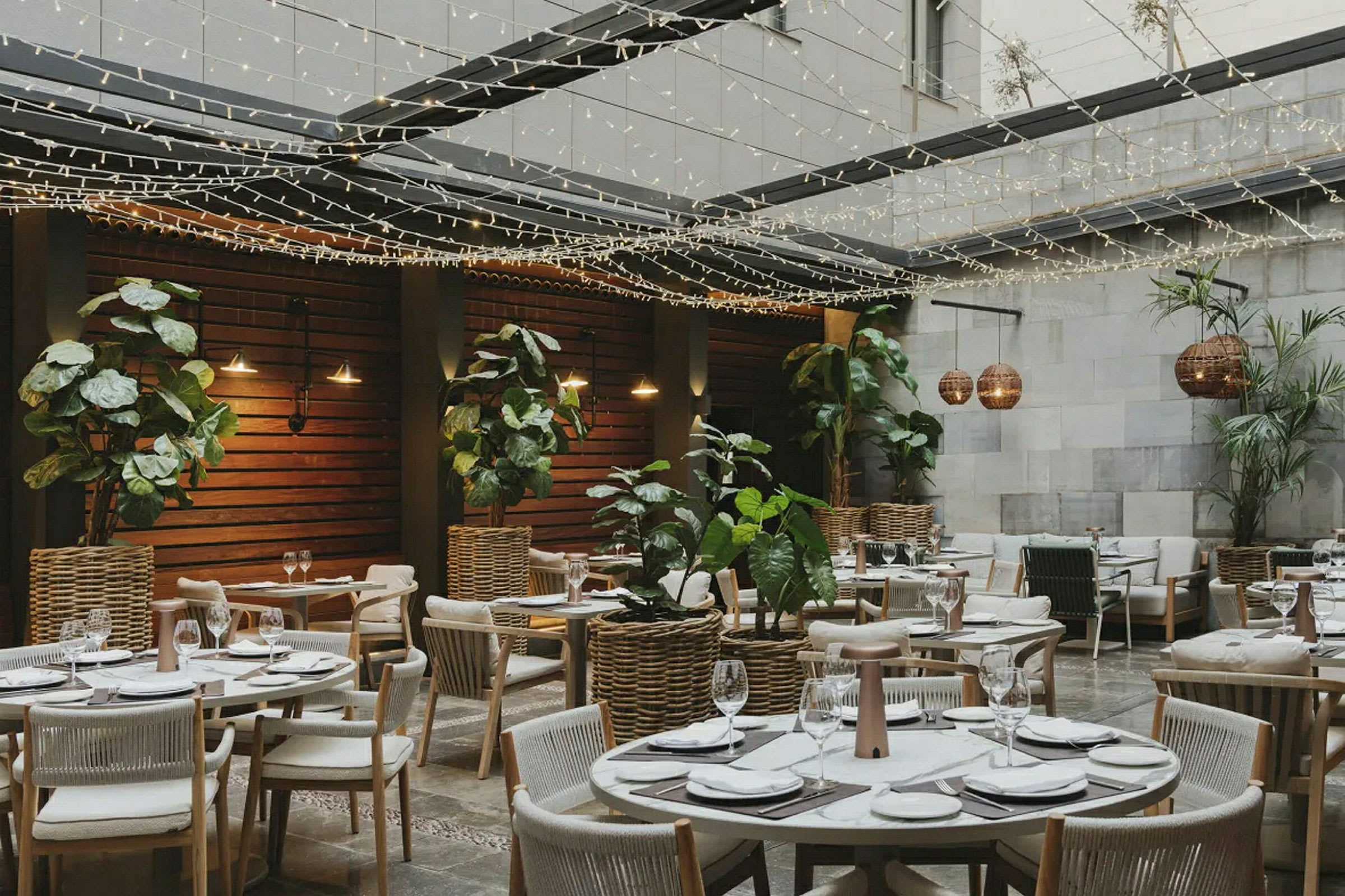 En restaurang med utomhusområde, bord dekorerade för måltider omgivna av växter och ljusslingor. (AI-genererad text)