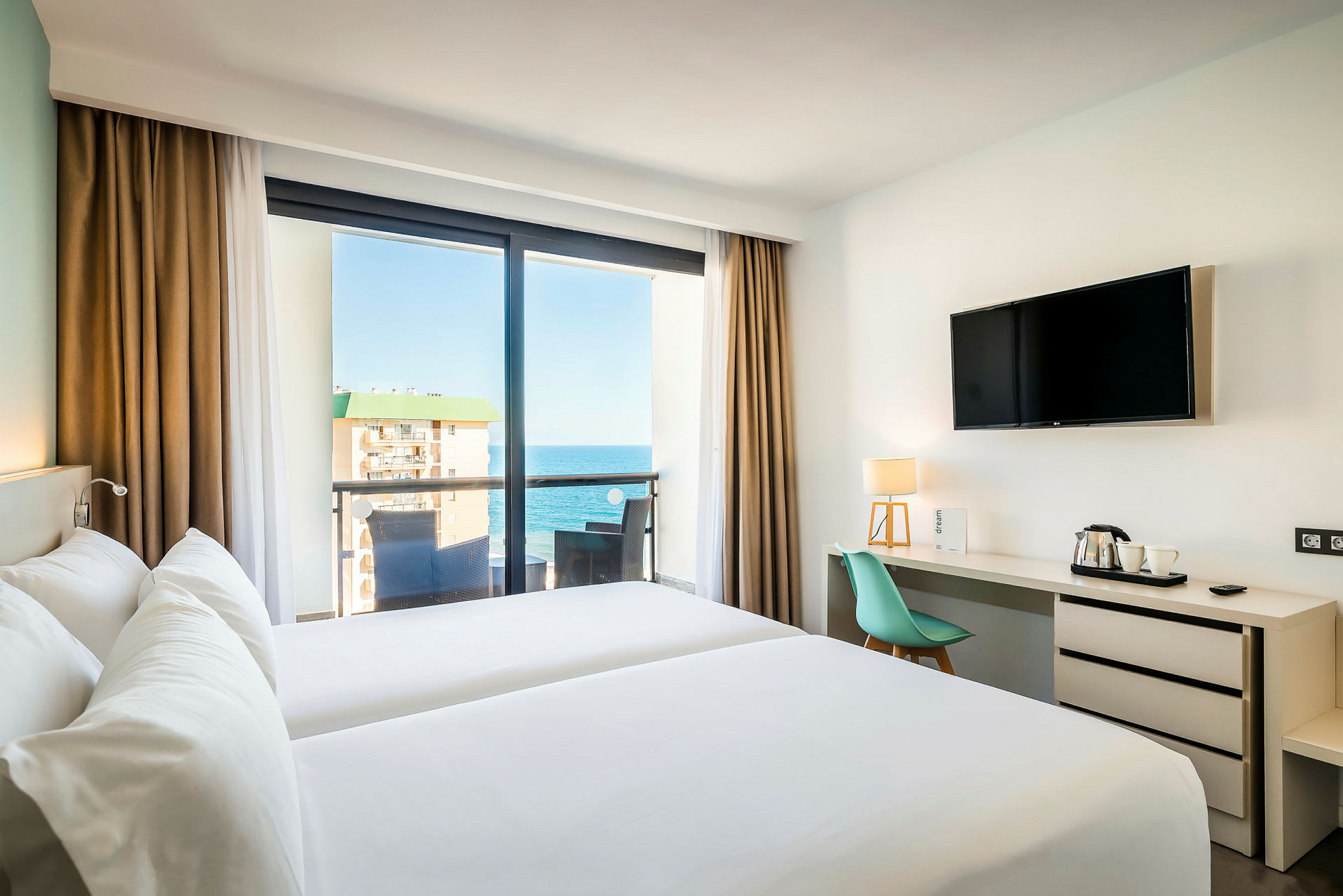 Ett hotellrum med två enkelsängar och en balkong med utsikt över havet. Rummet har även en TV och ett skrivbord med en stol och lampor. (AI-genererad text)