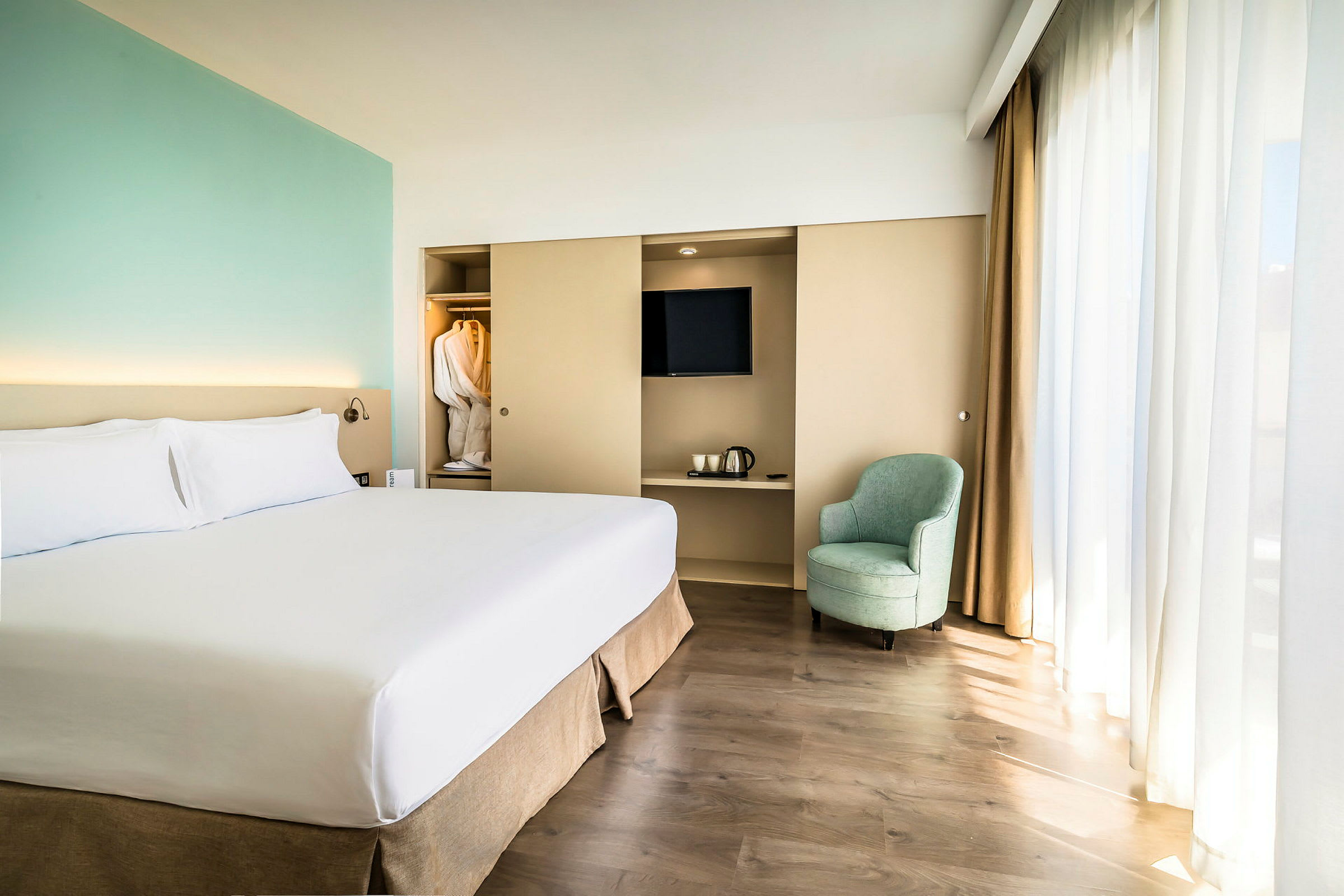 Ett hotellrum med en stor säng, en fåtölj, ett skåp med TV och en vattenkokare, samt en garderob med morgonrockar. (AI-genererad text)