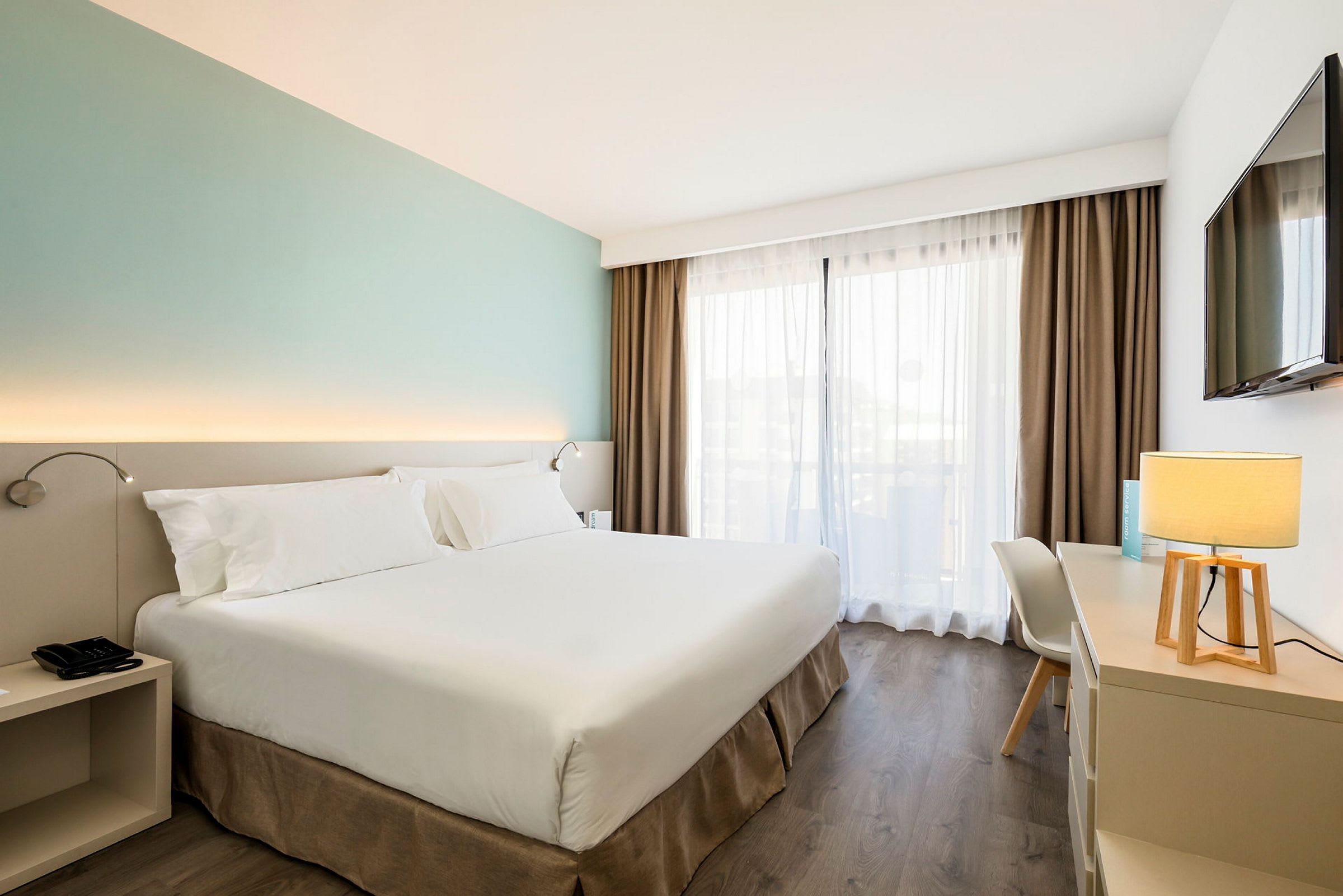 Ett rymligt hotellrum med en stor säng, skrivbord med en lampa och ett fönster med ljusgenomsläppliga gardiner. (AI-genererad text)