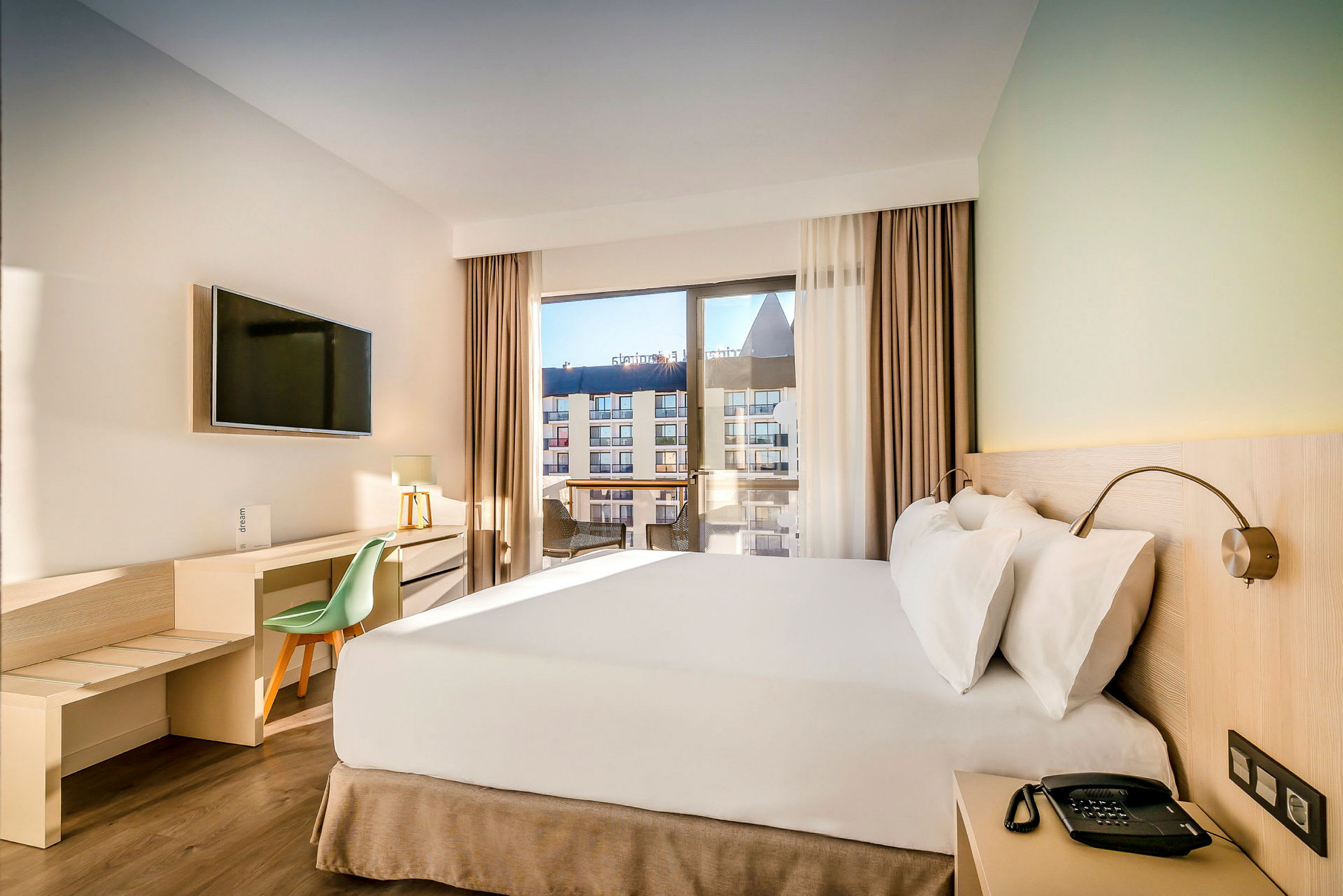 Ett hotellrum med en dubbelsäng, skrivbord och stor fönster med utsikt mot andra byggnader. (AI-genererad text)