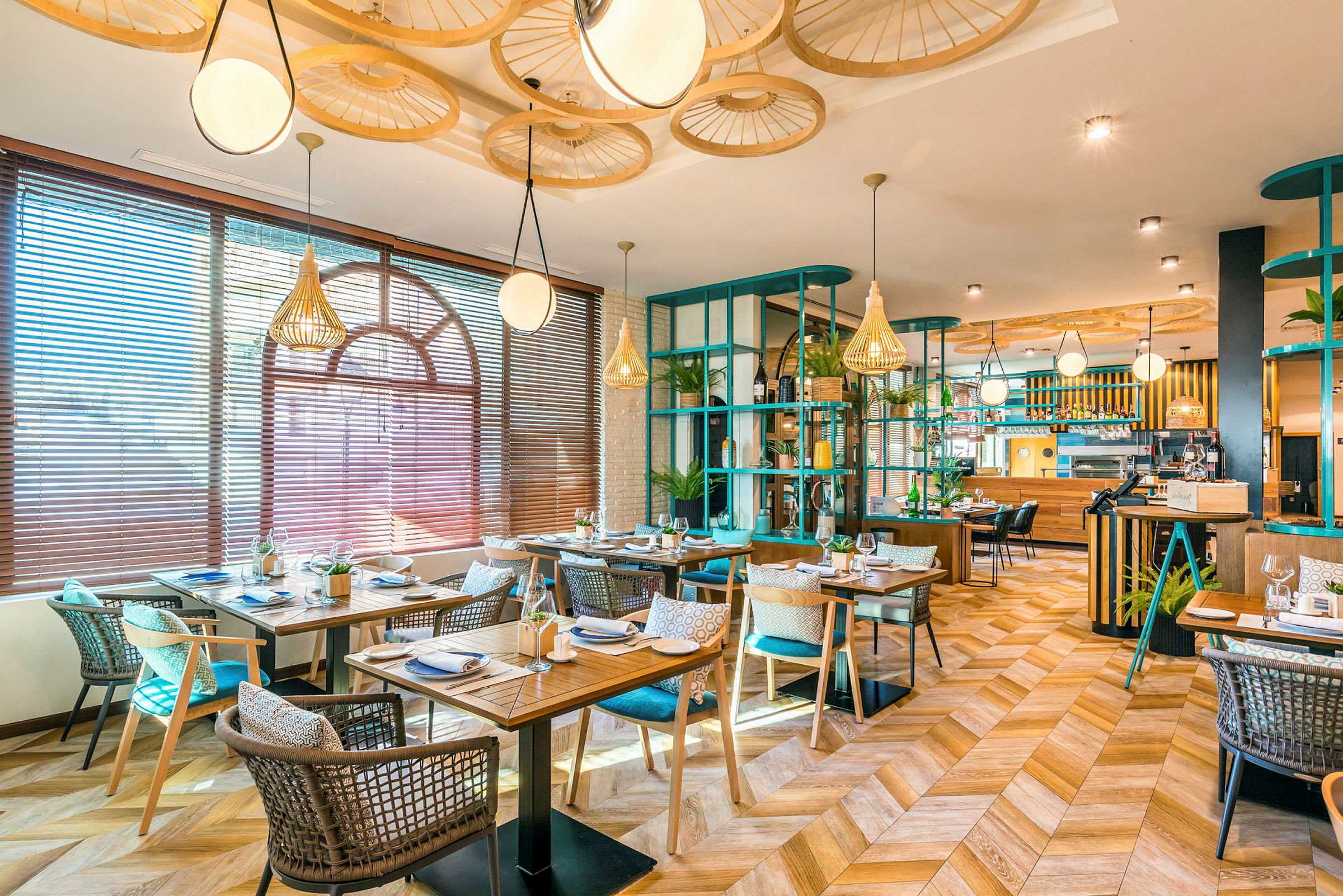 Ett modernt restaurangområde med träbord och stolar, dekorativa element i taket och stora fönster med persienner som släpper in naturligt ljus. (AI-genererad text)
