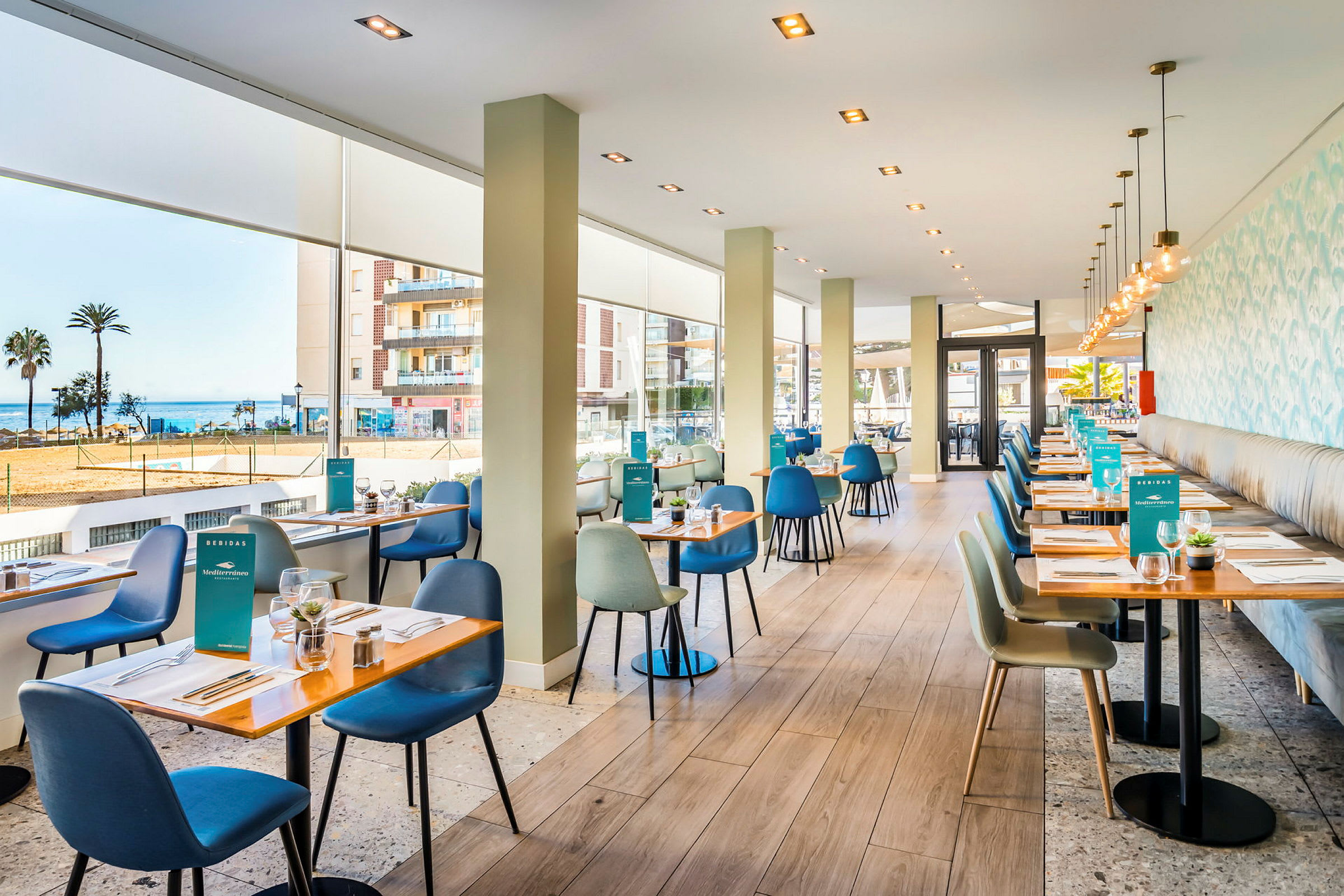 En fräsch och modern restaurang med stora fönster som visar utsikt mot havet. På borden finns dukningar och stolar i olika färger. (AI-genererad text)
