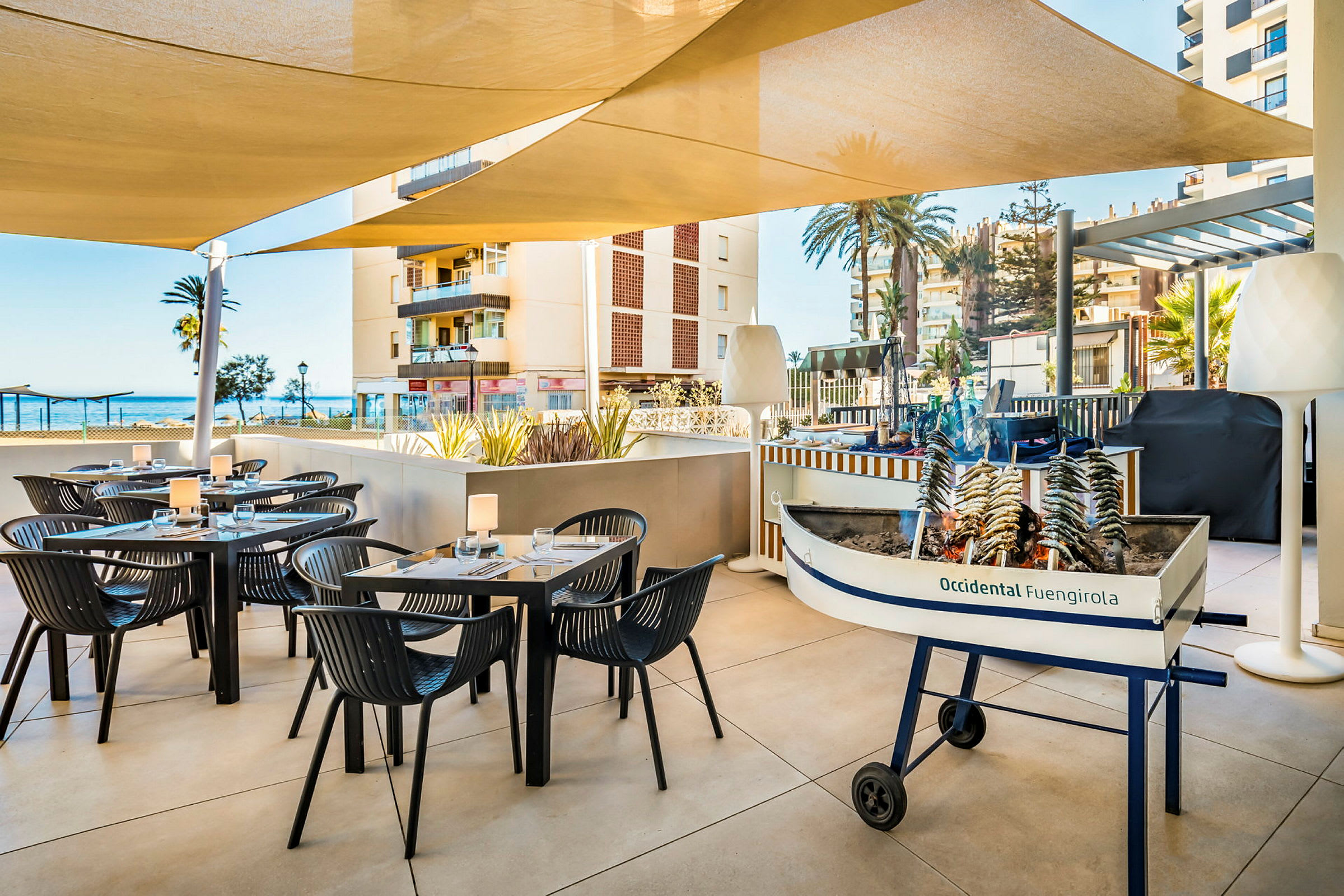 En uteservering vid en restaurang med bord och stolar under parasoller. I förgrunden finns en liten båtformad grill med sardiner på spett. Bakgrunden visar byggnader och havet. (AI-genererad text)