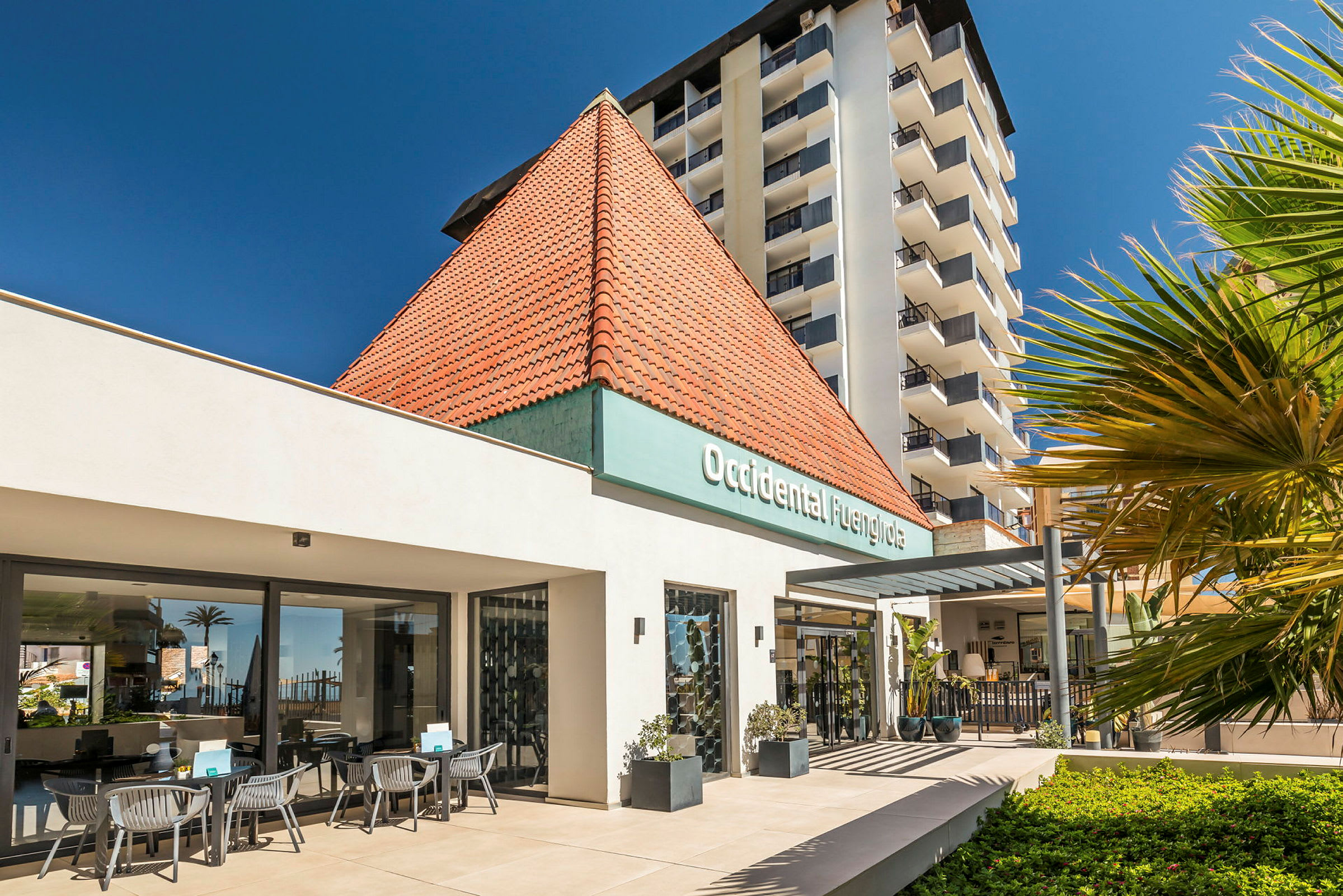 En modern hotellbyggnad med en ljus fasad och en karakteristisk rött tegeltak, omgiven av grönska och med uteservering. (AI-genererad text)