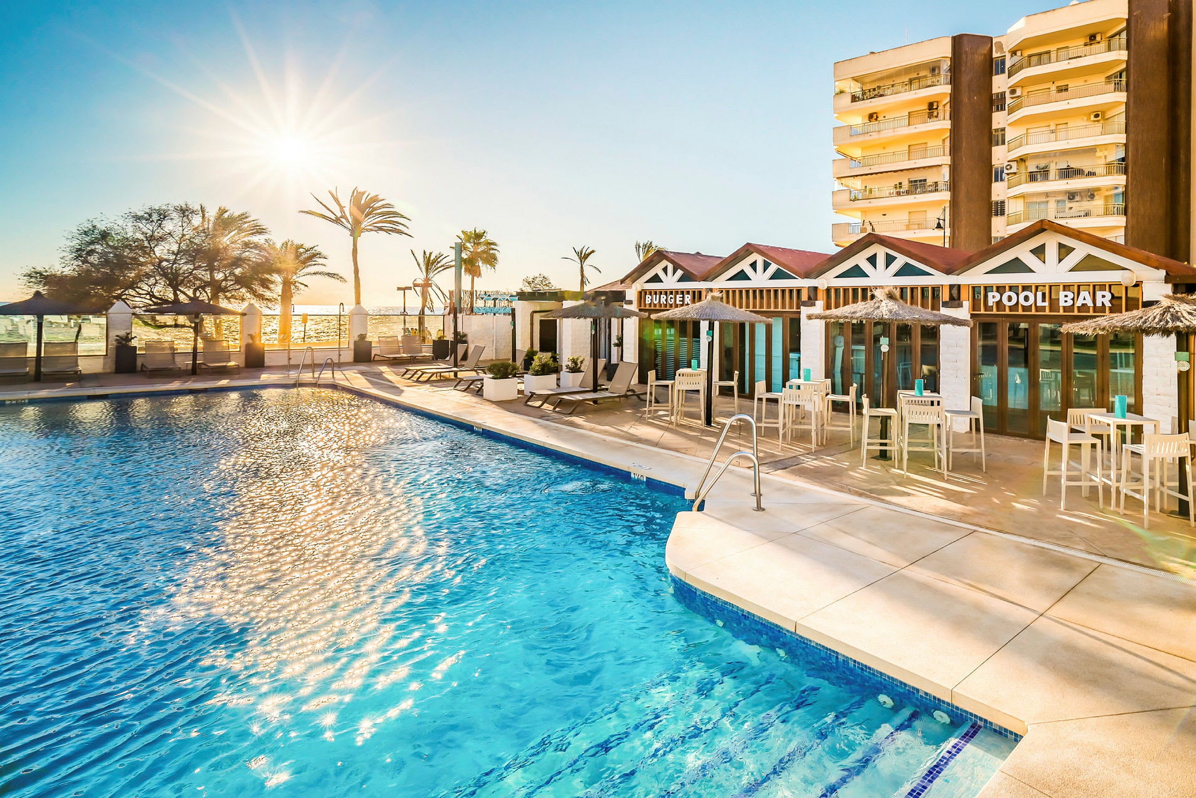 En swimmingpool vid solnedgång med solstolar och en restaurang med skyltar för "Burger" och "Pool Bar". I bakgrunden syns palmer och en byggnad. (AI-genererad text)